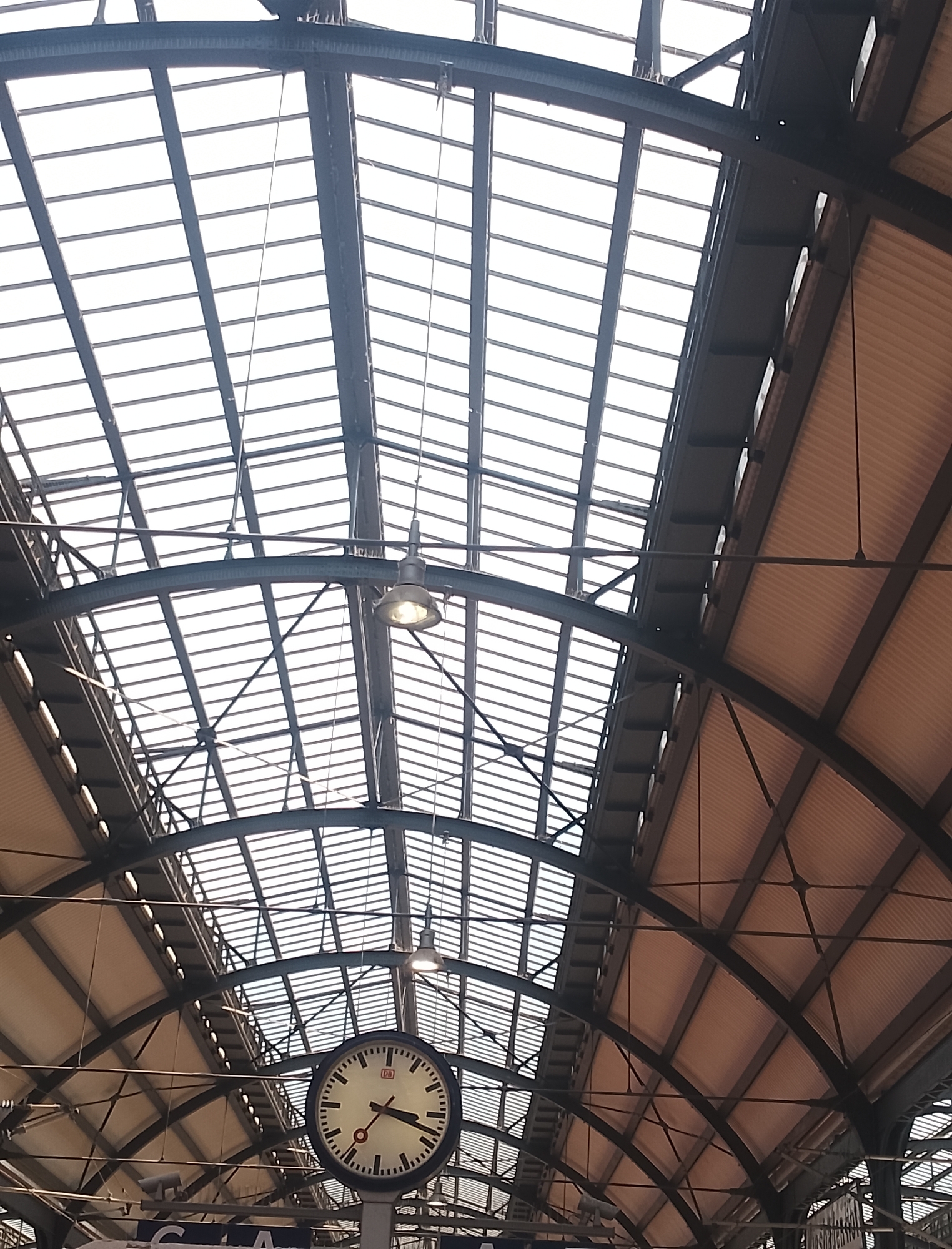An indoor photograph captures the interior of a train station's arched, ribbed roof structure. The ceiling is made of glass panels supported by a dark, angular metal framework. In the lower right quadrant of the image, a large, circular analog clock with a white face is prominently displayed. The clock has black hour and minute markers, a red second hand, and the "DB" logo, indicating it is a Deutsche Bahn clock. The clock is mounted on a pole. The overall scene depicts an architectural view of a spacious, well-lit train station, emphasizing its functional and aesthetic design.