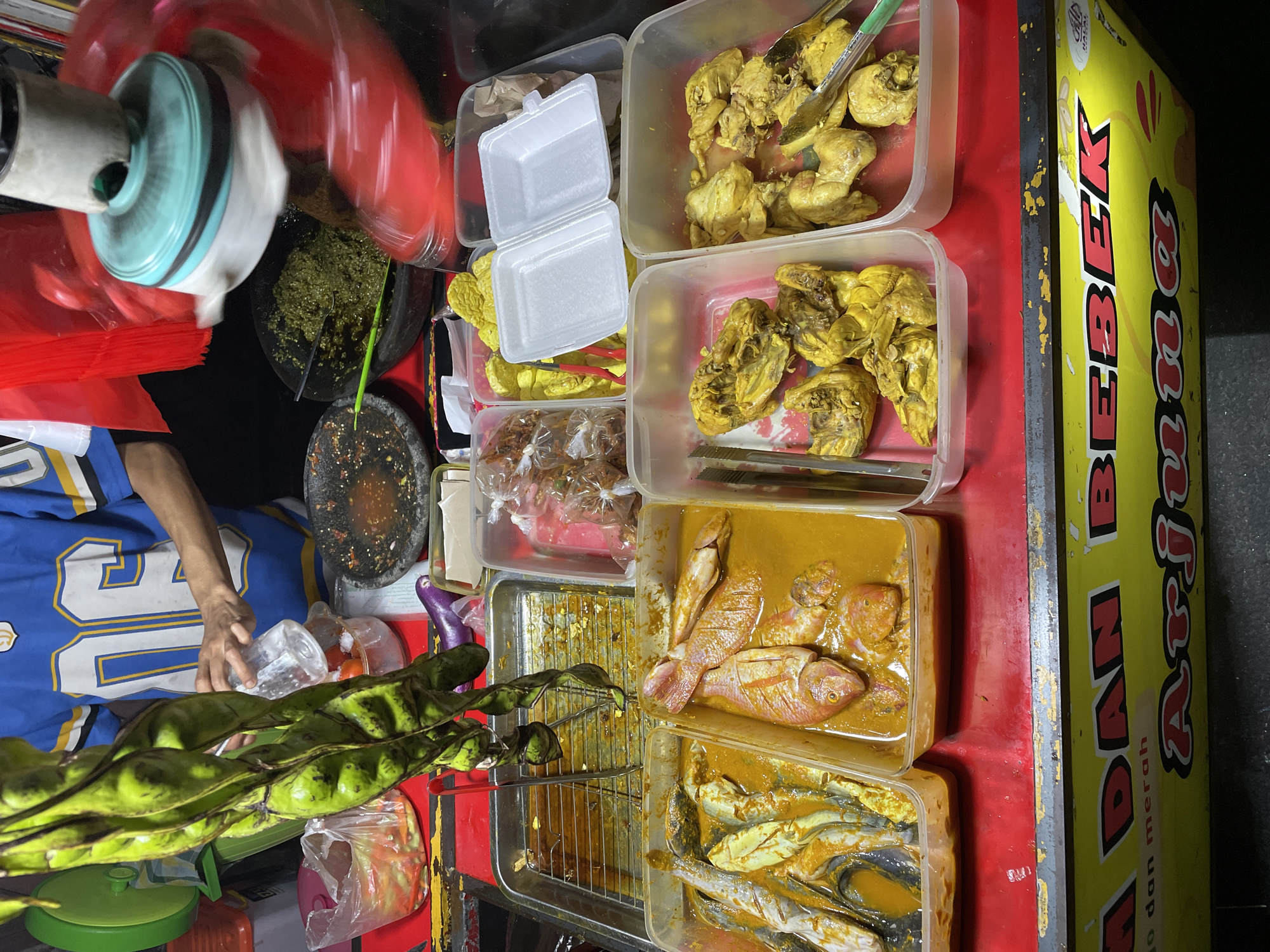 The image shows a street food stall in North Jakarta, Indonesia. The stall is set up with a variety of food items displayed in plastic containers. In the foreground, a person is wearing a blue and yellow jersey, with their arm visible as they interact with the food. Several bundles of green, long pods, likely petai, are prominently displayed.
The food includes several containers of what appears to be fried chicken, seasoned and cooked, some pieces with tongs. There are also containers of raw fish, possibly marinated in a yellow sauce, one with whole fish and another with smaller fish. A metal tray with a grill is also visible, suggesting further cooking or display. Other items include a large mortar and pestle with green paste, likely a sambal or seasoning, and some smaller plastic bags containing food items. A yellow sign with red lettering, partially visible, appears to be advertising "BEBEK ARJUNA," which likely indicates duck dishes are a specialty.
The scene is likely taking place outdoors, given the context of a street food stall. The lighting suggests it could be evening or night, but the weather is not discernible. The overall impression is one of a bustling food market or roadside eatery. The focus is on the colorful food presentation and the preparation or serving of meals. The visible text, "BEBEK ARJUNA," is a key detail indicating the type of cuisine offered.