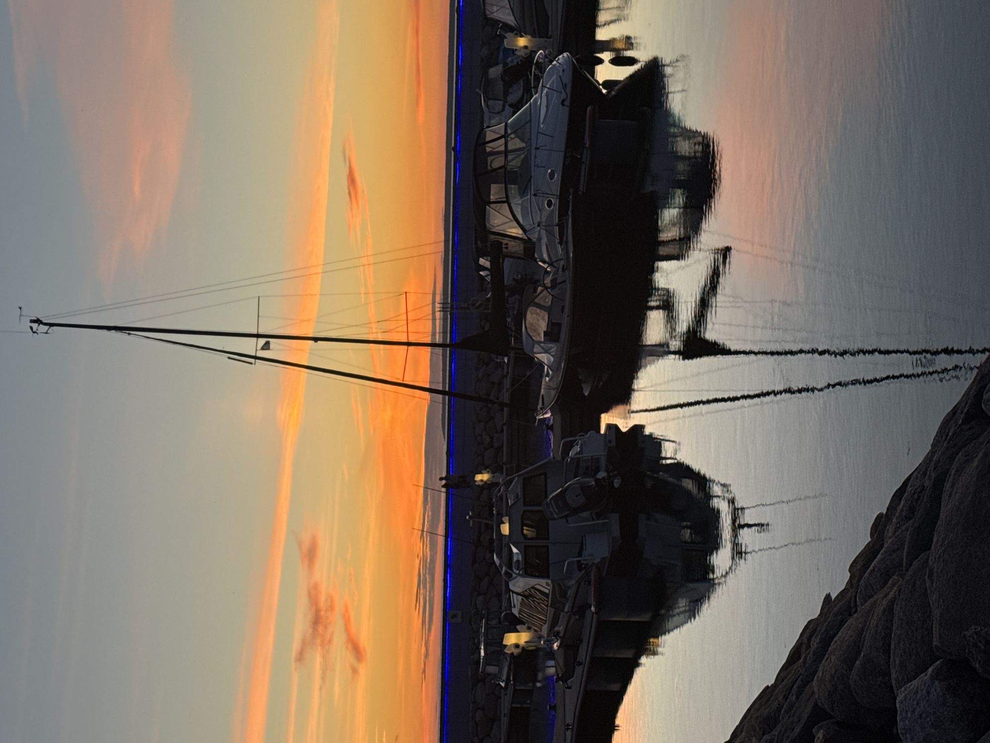 This is a vertical, full-shot photograph taken at sunset in Tallinn, Estonia, likely during the summer given the clear skies and calm water. The scene depicts a marina with several boats docked and their reflections shimmering on the still water. In the foreground, the mast and rigging of a sailboat stretch upwards towards a sky painted with soft orange and pink hues from the setting sun.
To the right, a modern cabin cruiser is prominently featured, its white hull and dark windows contrasting with the vibrant sky. The boat appears to be in pristine condition, possibly ready for a voyage or recently returned to shore. Its reflection is clearly visible in the water, creating a mirror image that emphasizes the tranquility of the moment. Further down the dock, another boat's outline is discernible, its form softened by the water's gentle ripples.
The background shows a stone embankment, likely part of the marina's infrastructure, and a line of blue lights bordering the dock, adding a touch of modern ambiance to the natural beauty of the sunset. The overall mood is peaceful and serene, capturing the quiet beauty of a maritime evening. There is no visible text in the image.