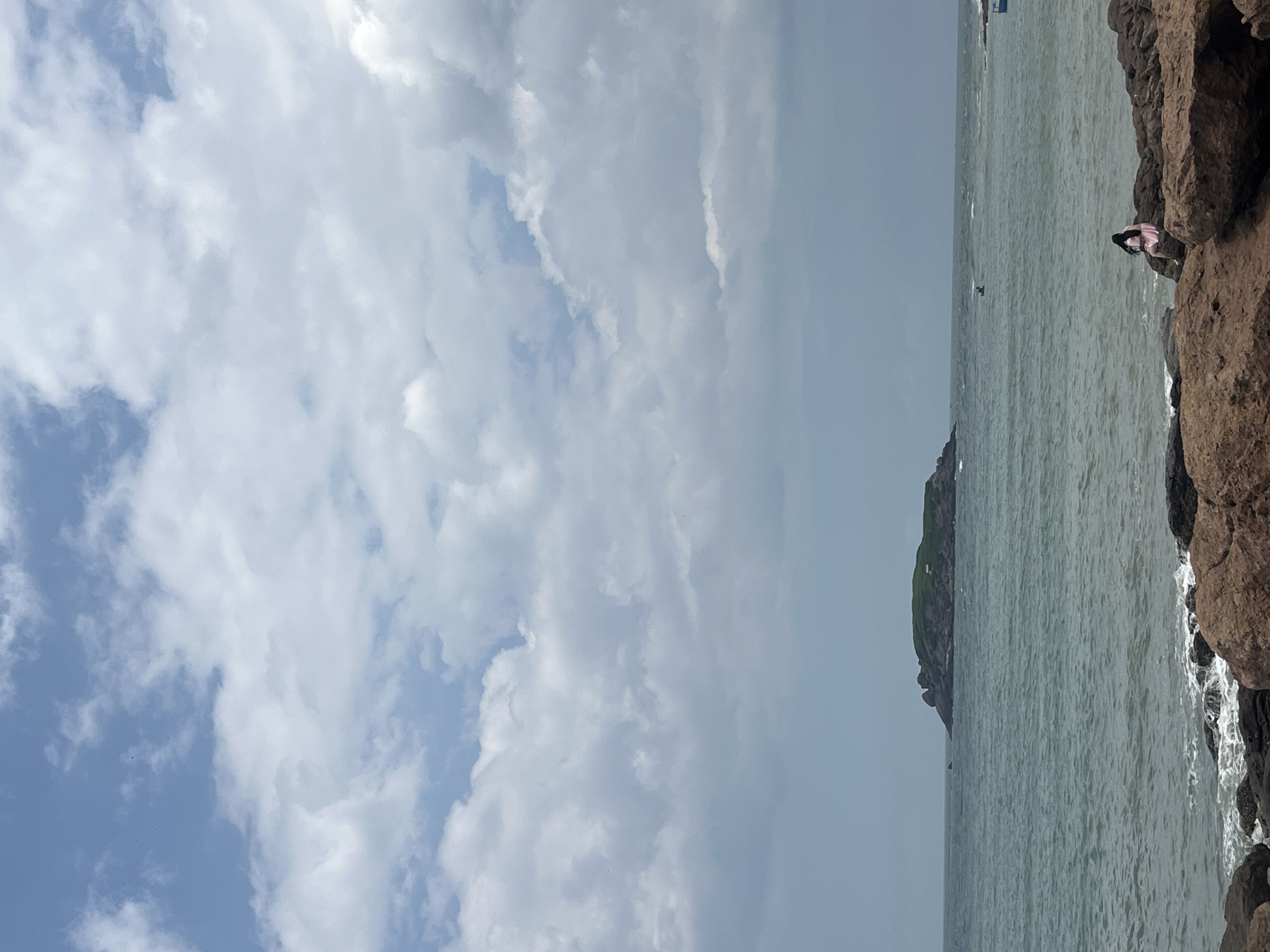 A wide shot shows a tranquil ocean scene with a small, green island in the distance under a cloudy sky. In the foreground, a person sits on large, reddish-brown rocks at the edge of the water. The sea is a deep teal with gentle waves lapping against the shore. The sky is filled with scattered white clouds against a pale blue background. The overall impression is one of natural beauty and quiet contemplation, likely captured at a beach location in Mui Ne, Vietnam.