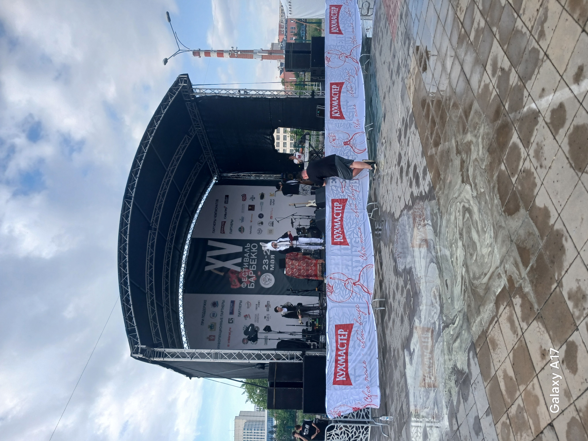 The image shows an outdoor stage set up for the "XV Festival Barbecue" event, held from May 23-26, as indicated on the backdrop. A banner reading "КУХМАСТЕР" (Kitchen Master) is displayed prominently. Musicians are visible on stage, and a person is seen leaning over a barrier. The ground is wet, suggesting a recent rain. In the background, buildings and a chimney can be seen, likely in Yekaterinburg, Russia, given the Cyrillic text and event name. The photo was taken with a "Galaxy A17" phone, as indicated by the watermark.