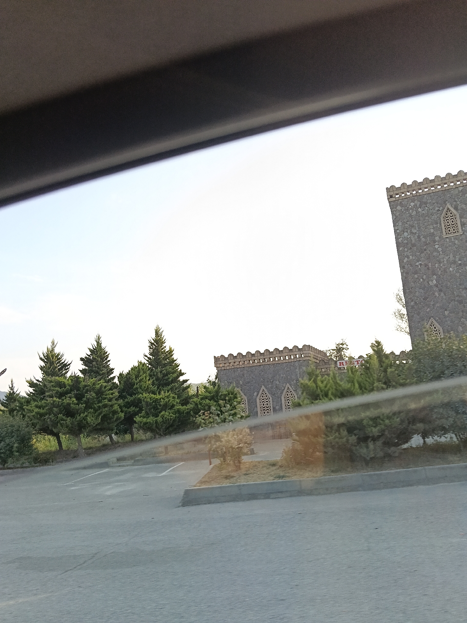 The image captures a view from inside a vehicle, looking out at a parking lot, a line of evergreen trees, and a building with a stone facade. The building appears to be made of rough-hewn stones and features decorative arched windows with intricate latticework. The top of the building has a crenellated parapet. In the background, a portion of another stone structure is visible, also with a crenellated top and decorative windows. A sign with the word "RESTO" in red letters is partially visible on one of the buildings. The sky is bright and overcast, suggesting it might be late afternoon or early morning. The parking lot is paved with asphalt and marked with white lines, indicating parking spaces. The trees are dense and green, with some sunlight filtering through their branches. The scene is set in the village of Vəlvələ, Azerbaijan, which is known for its historical sites and natural beauty.