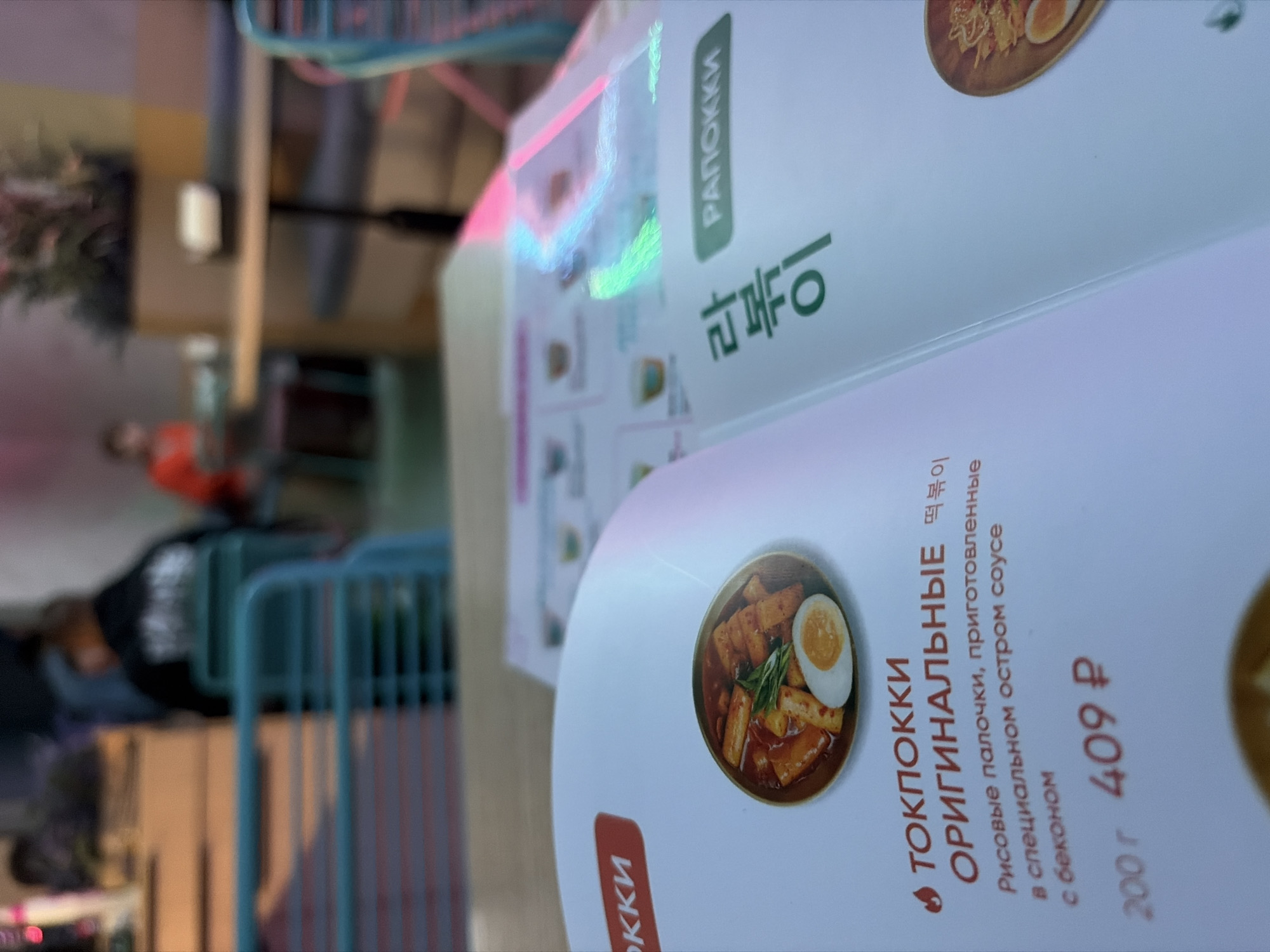 This image captures a detailed, close-up view of an open menu at a Chiko Korean bistro in Izhevsk, Russia. The foreground is dominated by the menu, specifically the left page featuring "ТОКПОККИ ОРИГИНАЛЬНЫЕ" (Tteokbokki Original). An illustrative photograph displays a bowl of tteokbokki, consisting of rice cakes, a half hard-boiled egg, and green garnish in a rich red sauce. The accompanying text describes it as "Рисовые палочки, приготовленные в специальном остром соусе с беконом" (Rice sticks, cooked in a special spicy sauce with bacon), priced at "200 г" (200 grams) for "409 ₽" (409 Russian Rubles). The adjacent right page of the menu shows Korean characters "라볶이" (Rabokki) and its transliteration "РАПОККИ" (Rapokki) against a green background. Distinct pink and green iridescent reflections from overhead lighting are visible across the glossy menu pages.
The background, out of focus, reveals the interior of a casual dining establishment. Several patrons are visible; a person in a dark top with a lighter graphic on the back is seated facing away from the camera in a light teal-colored chair on the left. Further to the right, a person wearing a red top is seated at a table. The furniture includes multiple light teal metal chairs and light-colored wooden tabletops. Dark grey upholstered bench seating is also discernible in the mid-distance. A large, blurred botanical arrangement with green foliage is positioned on a raised surface in the background, adding to the decor. The overall environment suggests a modern, vibrant eatery with ample lighting.