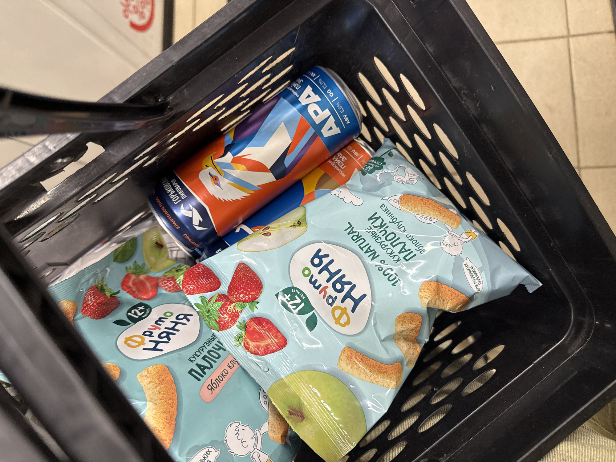 This is a close-up shot taken from above into a black plastic shopping basket. The basket contains two packages of "Fruto Nyanya" (Фруто Няня) corn sticks, apple and strawberry flavored, marketed for toddlers 12+ months. Alongside the snacks is a can of "Gorkovskaya Pivovarnya" (Горьковская Пивоварня) APA beer in blue, orange, and white packaging. There are no people visible. The location is likely a store in Russia based on the product packaging, but the precise city (including Moscow) cannot be verified from the provided image.