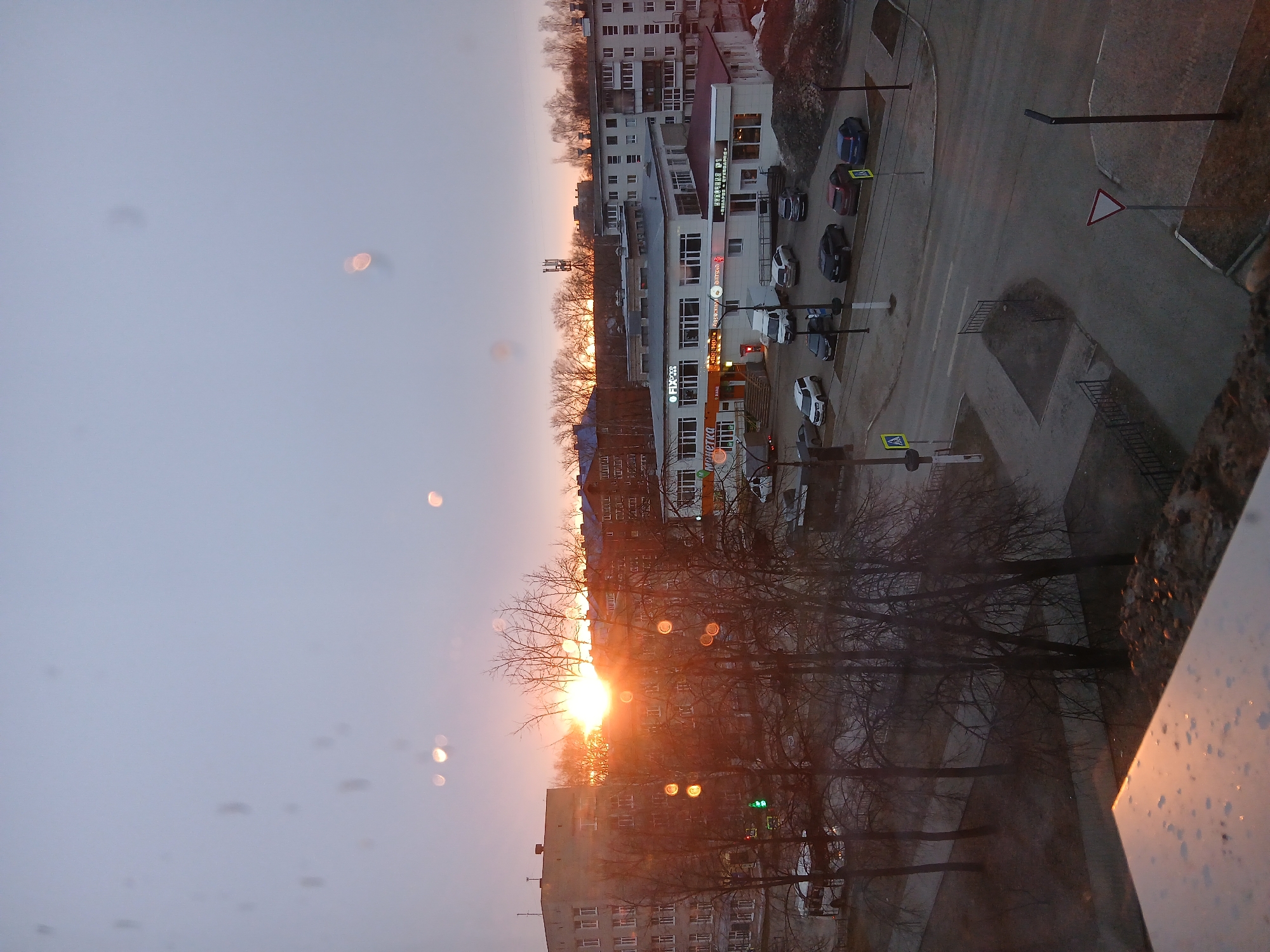 The image depicts a cityscape at sunset in Chaikovsky, Russia, observed from an elevated vantage point, likely a window. The sun is low on the horizon, casting a warm, orange glow through bare trees and onto buildings.
Buildings: Primarily residential apartment blocks are visible, along with a commercial building with the Cyrillic word "Пятерочка" on its facade, indicating a Petyorochka supermarket.
Road and Traffic: A street intersection is visible with parked cars and traffic lights.
Trees: Bare trees create a silhouette against the sunset.
Weather: The sky is overcast with a hint of orange from the setting sun. There are water spots on the window, suggesting recent rain or condensation.
Signs: Road signs, including a yield sign, are visible at the intersection.