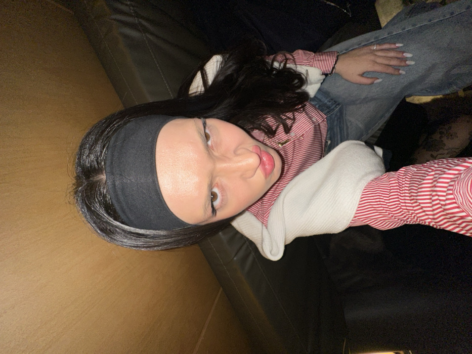 A young woman with dark hair and a black headband poses for a selfie while lounging on a dark leather couch. She wears a white sweater over a red and white striped shirt and blue jeans. Her face is slightly tilted with a pouty expression. Her manicured hand, adorned with a ring, rests on her leg. A dark shadow falls on the right side of the frame, with a wooden-paneled wall visible on the left.