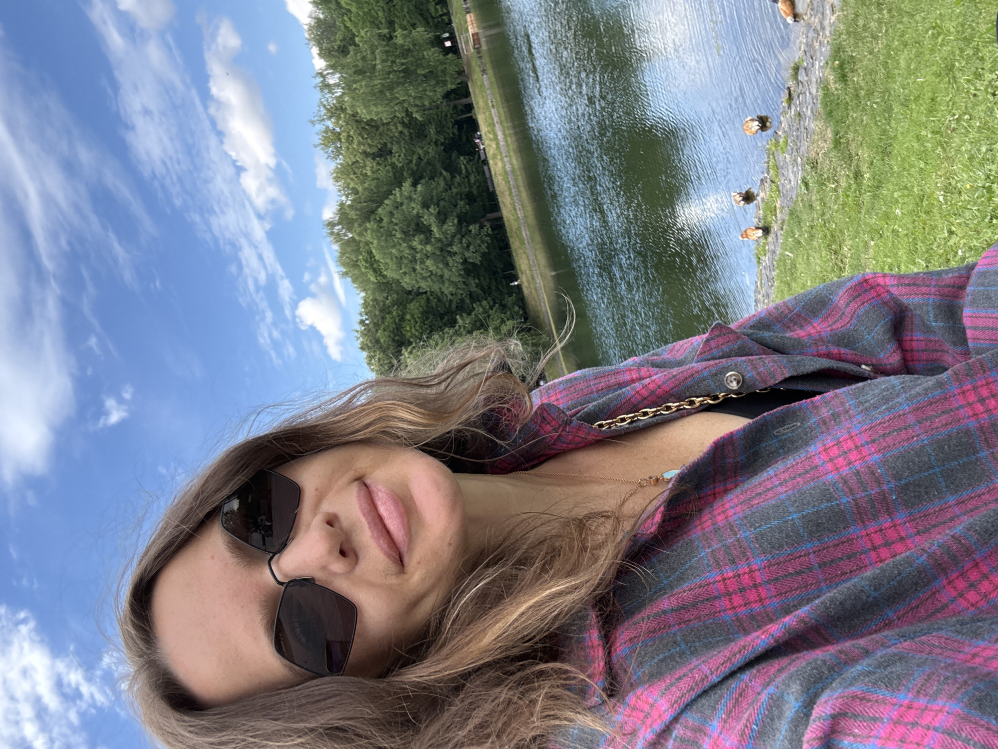 A woman with brown hair, wearing sunglasses and a pink and gray plaid shirt, takes a selfie by a lake. In the background, trees line the shore of the lake, and several ducks are visible in the water. The sky is blue with white clouds. This scene is likely in the city of Moscow, Russia, given the context provided by the prompt.