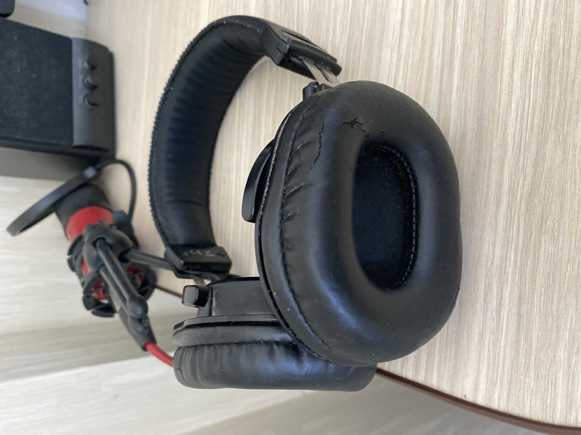 In the image, a pair of black over-ear headphones is positioned on a light-colored wooden surface, likely a desk or table. The headphones are resting with one earcup prominently in the foreground, showing some signs of wear, including minor cracks in the faux leather padding. Behind the headphones, a red and black microphone on a stand is visible, along with the edge of a black speaker. The background consists of a white vertical surface, possibly a wall or blind, and a wooden partition. The lighting suggests an indoor environment. There are no people or animals present, and no specific indication of the exact location within Chișinău, Moldova, beyond what can be inferred from typical household or office settings.