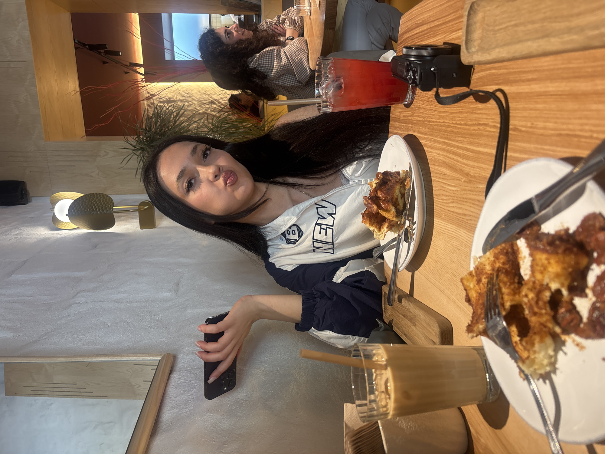 A woman with long, dark hair is pictured at a table, making a pouting face at the camera. She wears a white and navy blue jacket. A plate of food sits in front of her, along with a drink in a glass. She is holding a phone in her left hand.
Another woman with curly hair is seated at the table in the background. Other items on the table include a camera, a red beverage in a glass, and another plate of food.
The location is possibly a cafe or restaurant, given the presence of food and drinks. The city is Yerevan, Armenia.
