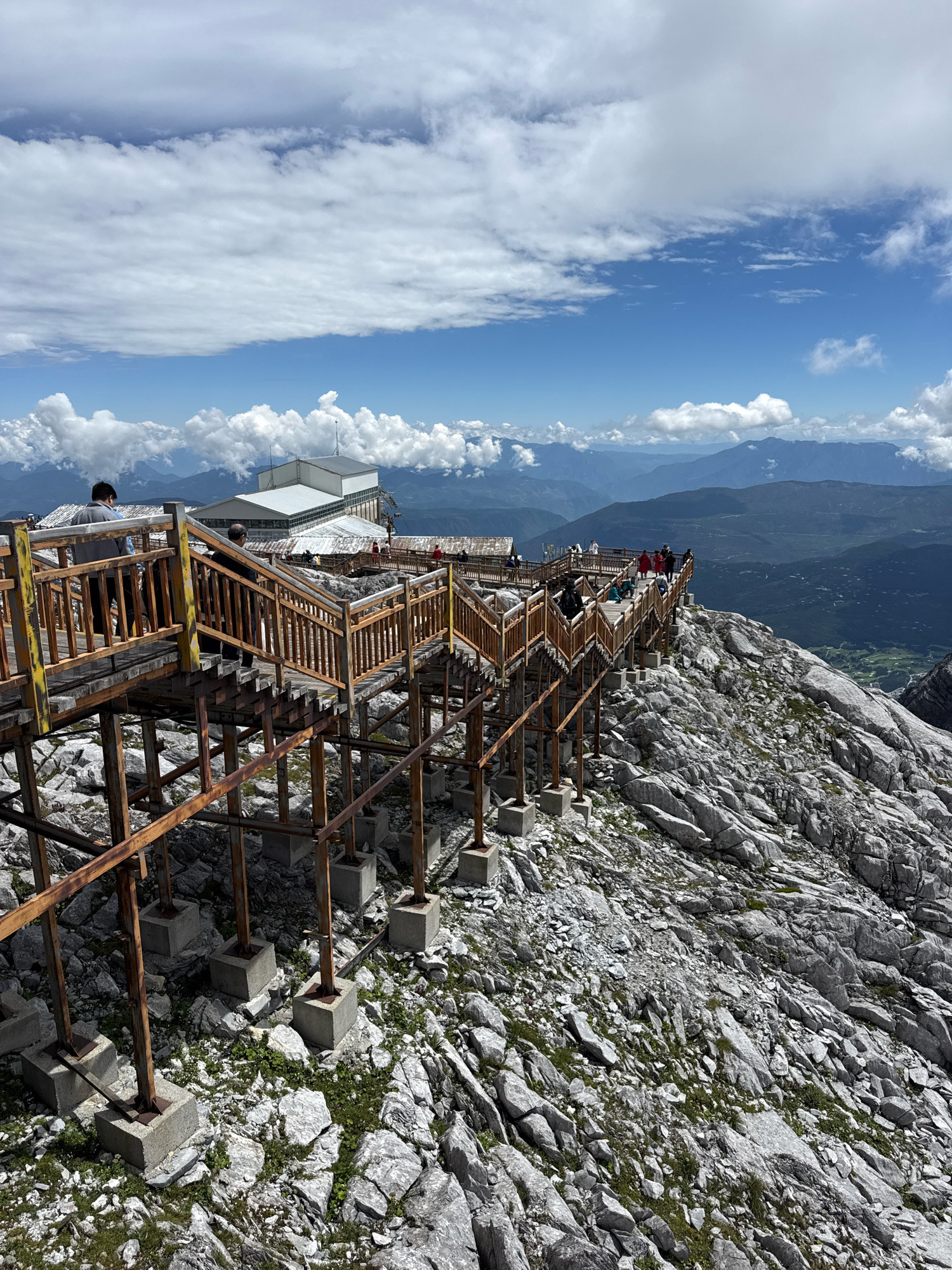 The image depicts a scenic viewpoint on a rocky mountain, likely located in Yulong Nakhi Autonomous County, China, given the context of the Jade Dragon Snow Mountain range, a popular tourist destination. A wooden boardwalk with stairs winds its way along the mountainside, offering visitors a path to traverse the rugged terrain and admire the panoramic views. Several people are visible on the boardwalk, some ascending and descending the stairs, while others are standing and observing the landscape. In the background, a modern building with a white roof is situated on the mountain, possibly a visitor center or a restaurant. The sky is partly cloudy, with fluffy white clouds against a bright blue backdrop, suggesting fair weather conditions. The surrounding environment is characterized by rocky slopes covered with sparse vegetation, and in the distance, layers of mountains recede into the horizon.