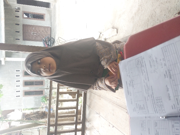 The image depicts a daytime scene in Lhokseumawe, Indonesia, observed from a low perspective. In the immediate foreground, a white paper document, appearing to be a form or worksheet with printed grids, text, and handwritten entries, lies open on a reddish-brown surface. A black pen with a silver clip rests on this document.
Centered in the midground, a young female child, estimated to be between 5-8 years old, is seated on a light-colored wooden floor or deck. She wears a plain brown hijab covering her head and chest, and a long-sleeved garment with a subtle pattern. Her hands are clasped together in her lap, resting on a yellow object, possibly a book or folder, with a dark green strap partially visible. Her gaze is directed upwards and to the right, away from the camera. To her left, a light-colored wooden railing structure is visible.
The background features two single-story buildings constructed of light grey concrete or stucco. The building behind the child displays three tall, narrow windows with brown frames and five small rectangular vents or decorative elements near the roofline. The building further to the right has a set of brown double doors, one similar window, and two rectangular vents. A black scooter (motorcycle) is parked in front of the rightmost building. The ground beyond the wooden floor appears to be a light-colored concrete or dirt surface. Sparse vegetation is visible on the far left edge of the frame.