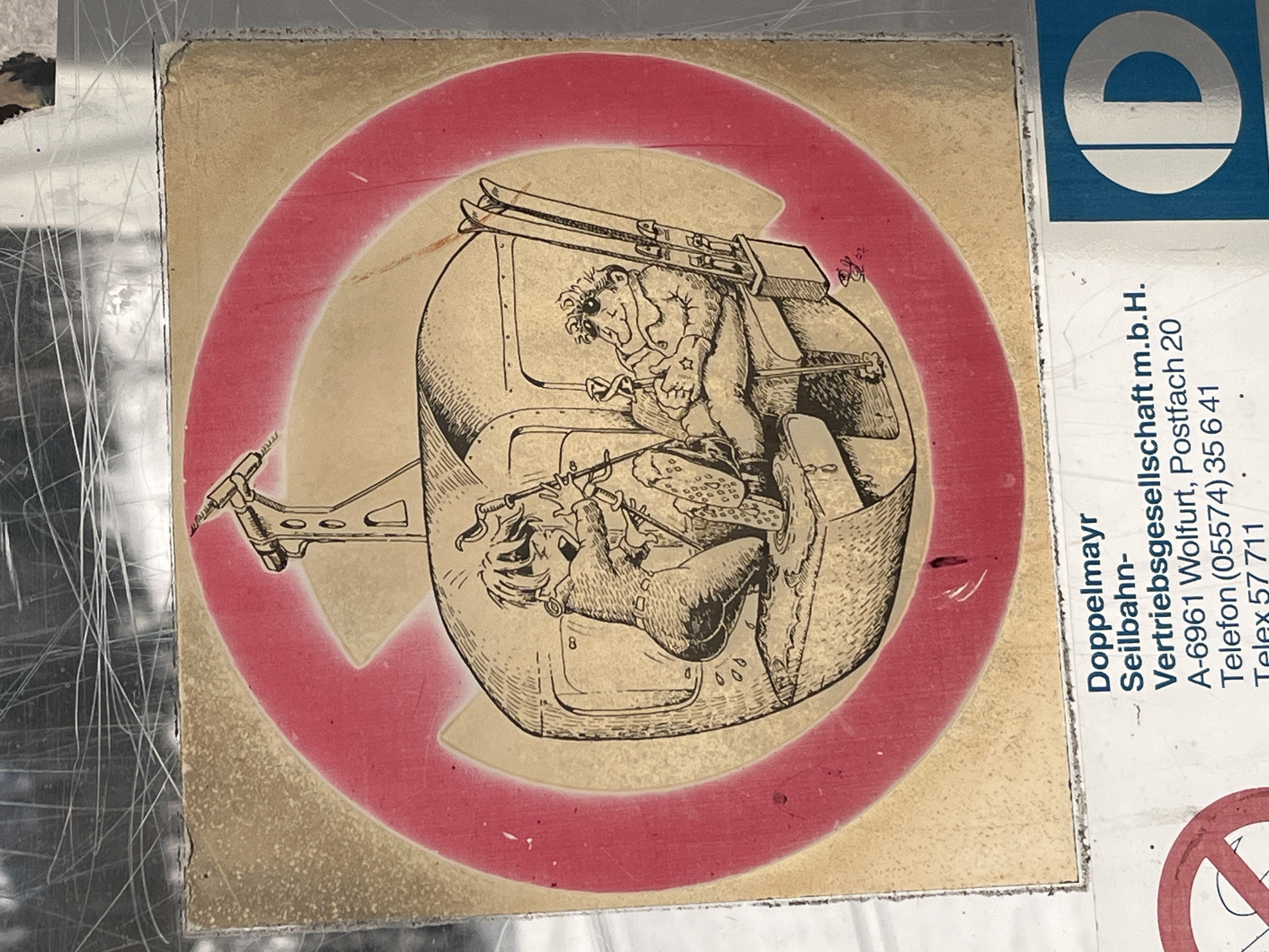 The image shows a sticker or sign, likely found in Wolfurt, Austria, as indicated by the text on the right side: "Doppelmayr Seilbahn-Vertriebsgesellschaft m.b.H. A-6961 Wolfurt, Postfach 20 Telefon (05574) 35 6 41 Telex 57 711." To the left of the text is a blue and white logo. Adjacent to this is a circular design, primarily tan with a broad red ring forming a prohibition symbol (a circle with a diagonal line through it). Inside the circle, a detailed illustration depicts two cartoon-like characters in a gondola-like structure. One character, presumably the operator, is on the upper level, wearing goggles and operating controls. The other is below, with what appears to be an animal or creature beside him. Skis are placed on top of the gondola. The overall image suggests a warning or prohibition related to the Doppelmayr Seilbahn company, involving the characters depicted. There is visible wear and tear on the sign. The background is blurred.