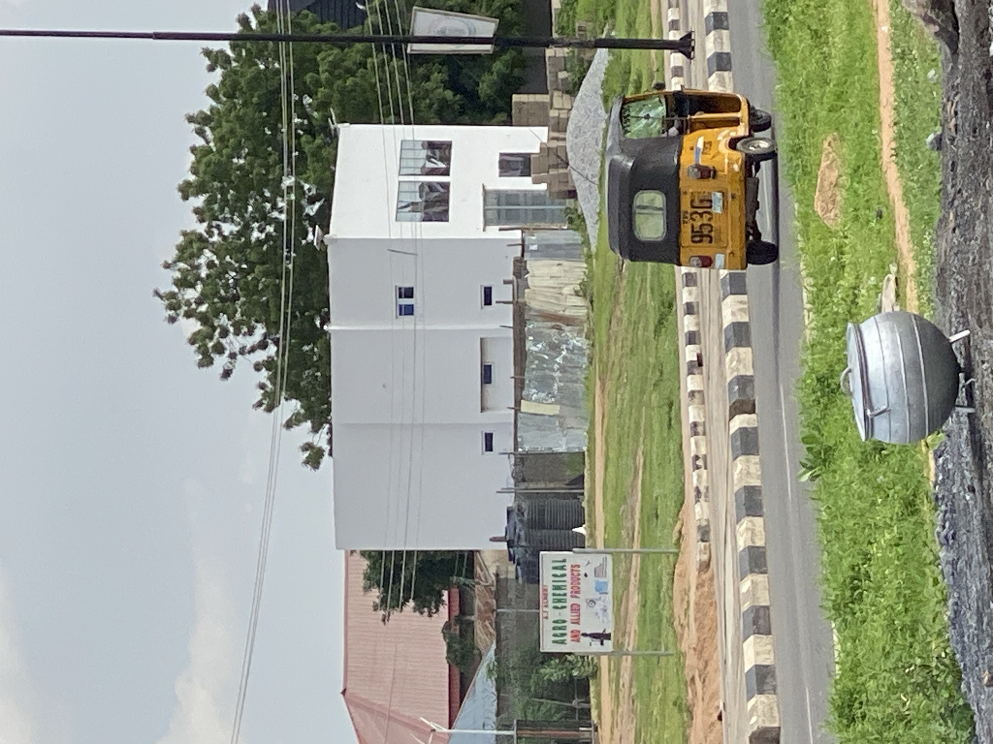 This image shows a street scene in Jalingo, Nigeria. A yellow and black auto-rickshaw is parked on the side of a road with a black and white striped curb. Behind the auto-rickshaw is a white building with several windows. Further back, there are more buildings and trees. A sign for "AGRO-CHEMICAL AND ALLIED PRODUCTS" is visible on the right side of the image. The sky is overcast, suggesting it could be a cloudy day. The scene appears to be in a semi-urban or rural area given the combination of buildings, roads, and vegetation. No people are clearly visible, but the presence of the auto-rickshaw suggests it's a populated area. The time of day is unclear due to the lighting.