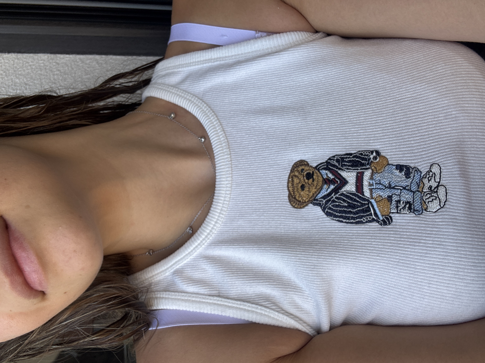 A woman with wet, dark hair is wearing a white ribbed tank top with a bear embroidered on it. She is also wearing a delicate silver necklace with small charms and a light purple bra. The image is a close-up shot of her upper body. The background appears to be an outdoor setting, possibly a wall with vertical slats. The lighting suggests it is daytime. The location is not specified in the provided information.