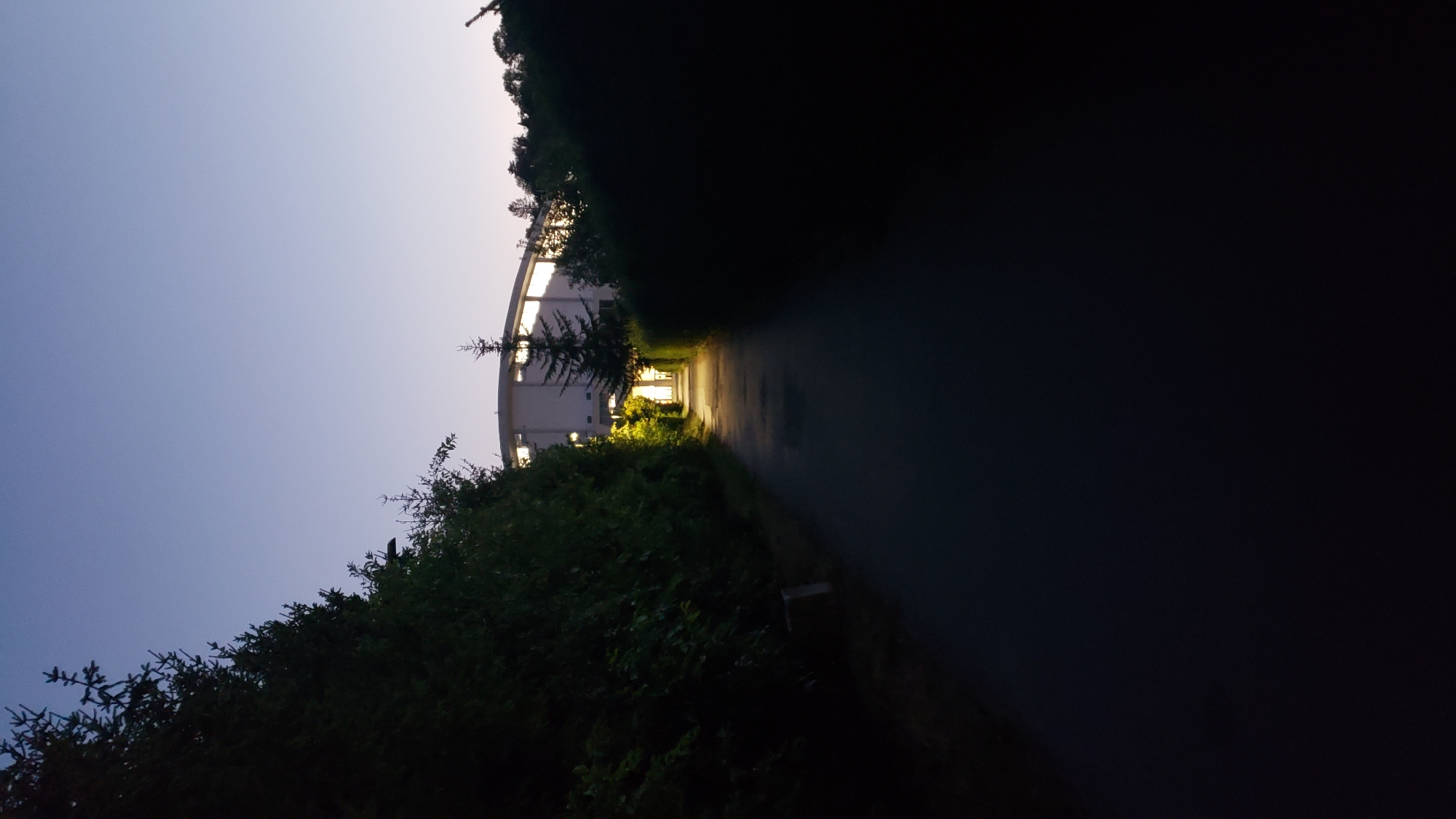 The image shows a pathway leading towards a brightly lit building at dusk, with trees and hedges on either side. The sky is a clear gradient of deep blue to light blue, indicating twilight. The pathway is dark asphalt and appears to be illuminated by lights from the building and possibly some pathway lights, casting a warm glow. The building itself is modern, with a curved facade and large windows that are brightly lit from within, suggesting activity. There are no people visible in the image. The scene suggests an entrance to a public or institutional building, such as a school, office, or community center, in an outdoor setting as night begins to fall. The location is the city of Prešov, Slovakia.