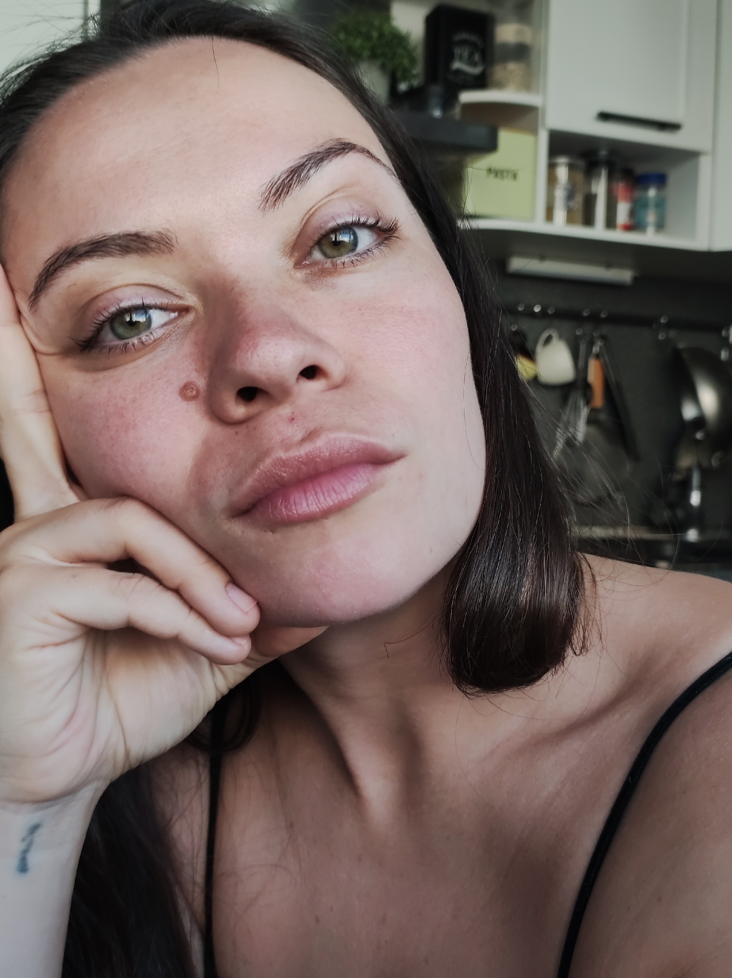 A close-up photograph shows a woman resting her face on her hand. The location is likely an interior kitchen setting.
The woman has dark hair and light green eyes. She has a mole on her left cheek near her nose. Her skin appears fair. She is wearing a thin-strapped top. On her left wrist, there is a small tattoo.
In the background, kitchen cabinets and cooking utensils are visible. The photograph is taken from a slightly low angle. The scene is domestic, and the lighting suggests it is daytime. The location in the city of Chaikovsky, Russia, cannot be confirmed from visual information alone.