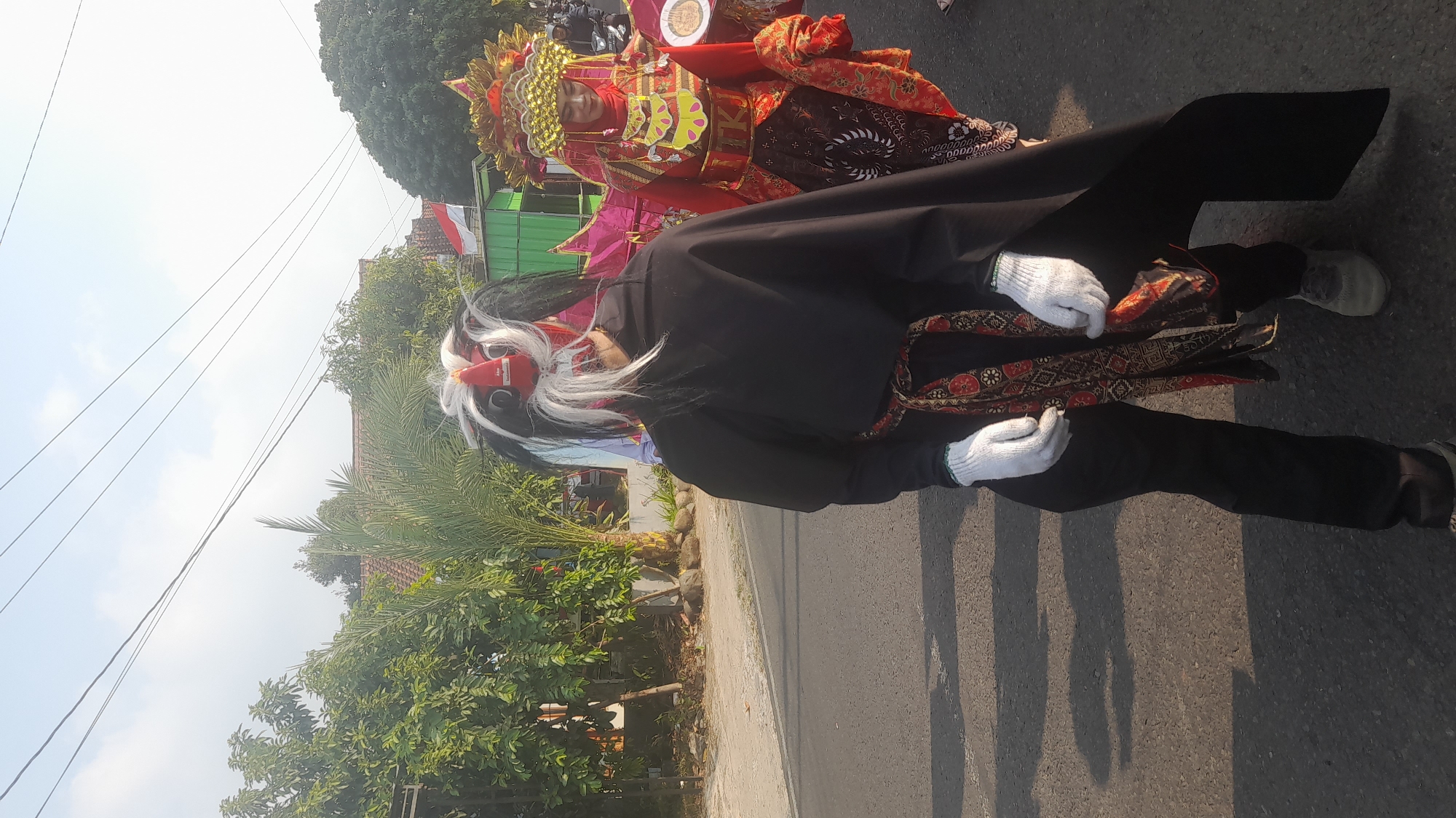 In Lemahabang, Indonesia, on a paved road with shadows cast by overhead power lines and trees lining the left side, two individuals in traditional attire participate in an outdoor event. In the foreground, a person wears a black cloak, black pants, and white gloves, with a patterned sash around their waist. Their head is adorned with a mask featuring white and black hair, and a red face with prominent eyes. To the right, a second person is dressed in a vibrant red and gold costume, including a decorative headdress. They are also wearing a red hijab. The background shows foliage, buildings, and parked motorcycles, suggesting a public gathering or procession. The sky is clear and bright.