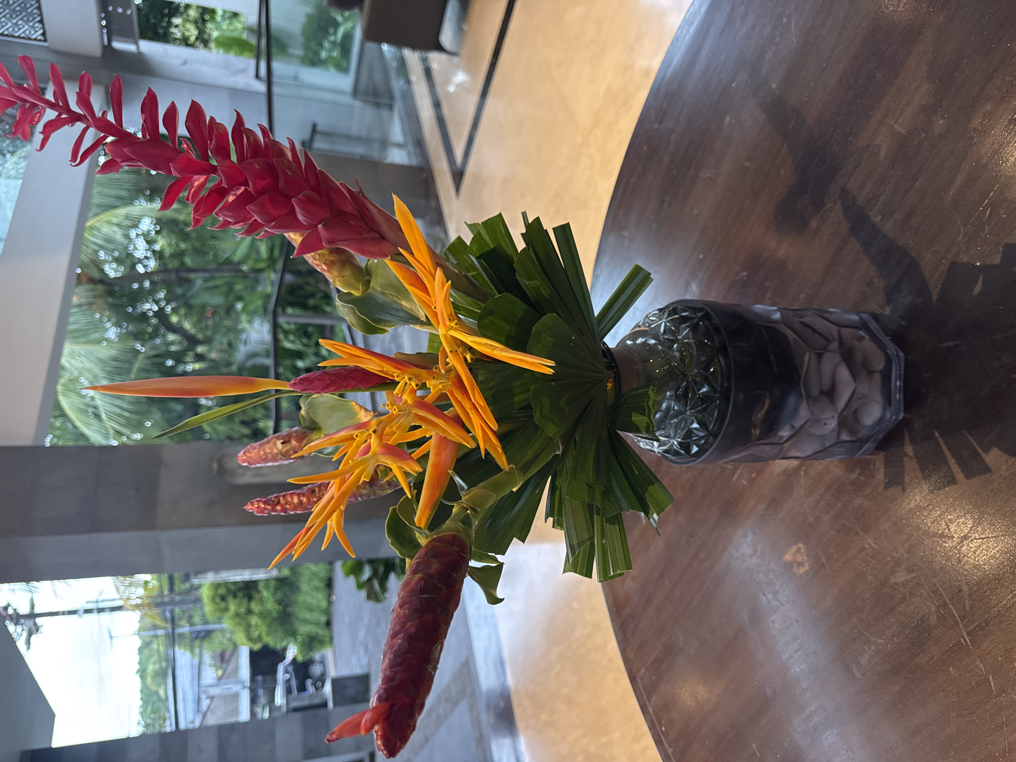 A vibrant tropical floral arrangement is displayed on a round, dark wooden table. The centerpiece features striking red ginger flowers, characterized by their elongated, torch-like inflorescences, alongside bright orange and yellow heliconia blooms, which resemble bird-of-paradise flowers. These colorful flowers are complemented by dark green, broad leaves arranged around their base, all held within a clear, textured glass vase. The wooden tabletop, positioned horizontally in the frame, exhibits a visible grain and minor surface wear.
In the background, filtered daylight illuminates an indoor space with light-colored flooring and sleek structural elements. Beyond these, large glass partitions reveal a lush outdoor environment dominated by tropical greenery, including discernible palm trees. Reflections of this verdant scenery are visible in the glass, creating a soft, blurred backdrop that hints at an expansive garden or natural landscape adjacent to the indoor area. A subtle shadow of the flower arrangement is cast upon the table. The overall scene suggests a decorative element in a sophisticated, possibly resort or hotel, setting in a tropical region like Kuta Selatan, Indonesia. No people are visible, and no specific activities are taking place. There is no visible text in the image.