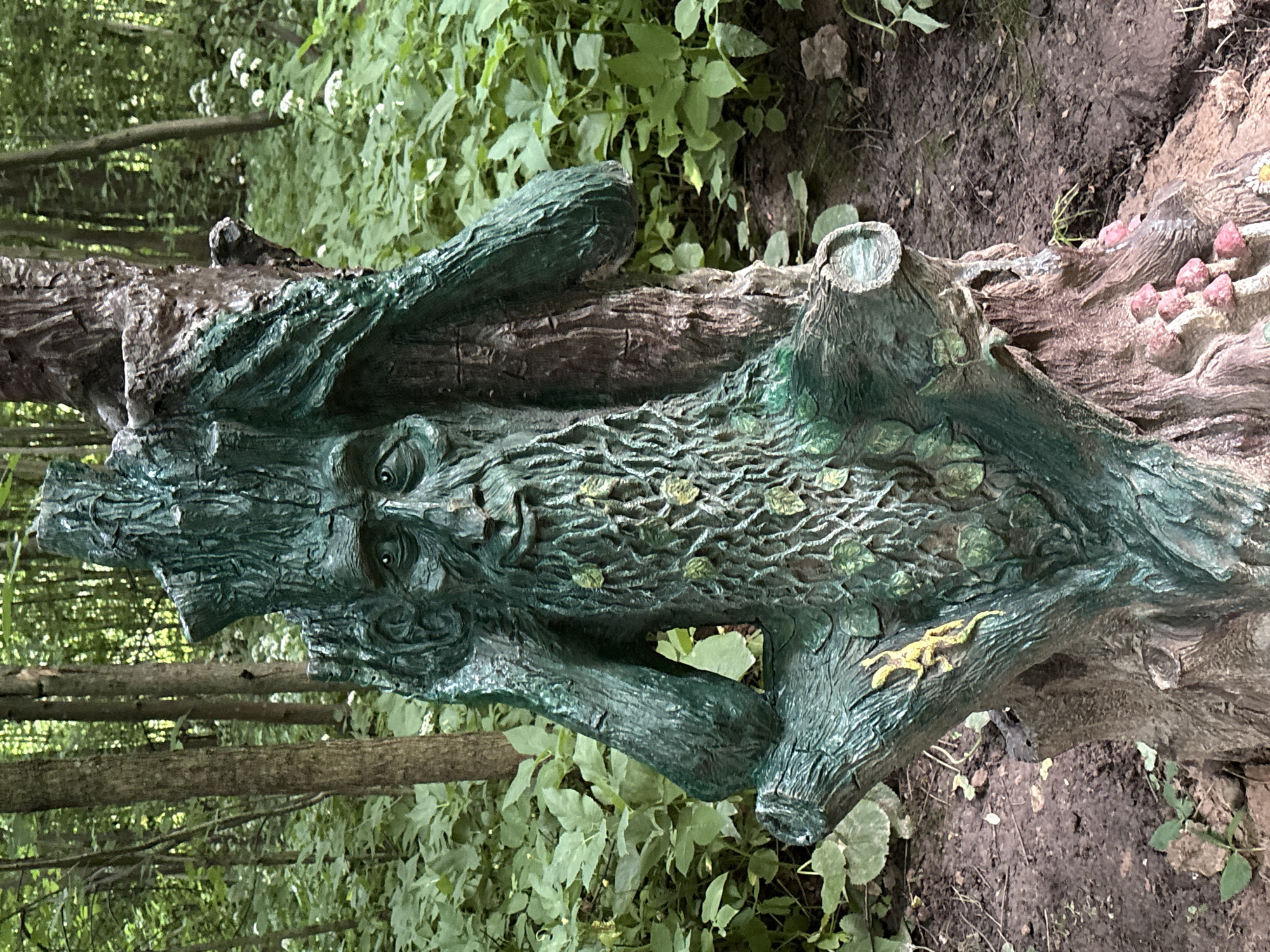 A sculpture of a green-faced, bearded tree spirit is situated in a forest setting. The sculpture is attached to a tree trunk and appears to be made of a weathered material, possibly concrete or resin, painted in shades of green and brown. The tree spirit has a long, textured beard adorned with small green leaves, and its hands are raised to its head as if in thought or distress. The background consists of lush green foliage, trees, and a muddy ground. Small pink mushroom figures are visible at the base of the sculpture. The location is identified as Горки, Russia.