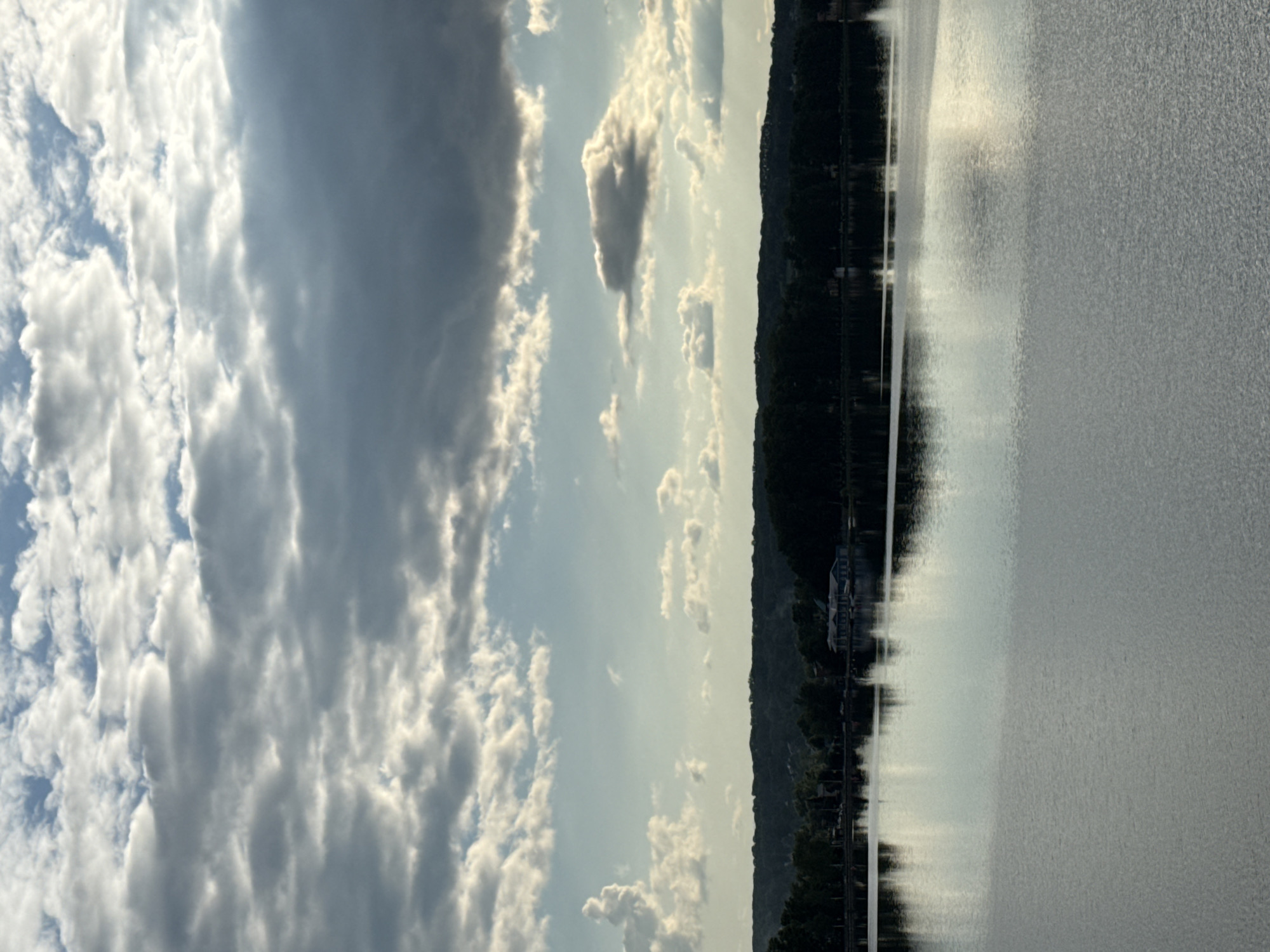 The image presents a tranquil, expansive landscape featuring a large body of water, likely the Votkinsk Reservoir in Votkinsk, Russia. The scene is dominated by a wide expanse of water in the foreground and midground, its surface covered in small ripples and reflecting the sky and distant shore. Bright sunlight glints across sections of the water, indicating a clear, sunny day despite the cloud cover.
A dark, dense treeline or hillside forms the distant shoreline, stretching horizontally across the image. Upon closer inspection, faint details of buildings, possibly houses or boathouses, are discernible nestled among the trees on this distant bank, with their reflections also visible in the water.
The upper half of the frame is filled with a dynamic sky, abundant with a mixture of cumulus and stratocumulus clouds. These clouds vary in tone from brilliant white, brightly lit by the sun, to darker shades of grey, suggesting varying thickness and shadow. Patches of clear blue sky are visible between the cloud formations.
The photograph captures the scene during the daytime, likely mid-morning or late afternoon, given the angle and intensity of the light reflecting off the water and illuminating the clouds. The weather appears fair with scattered clouds. There are no people or discernible text visible in the image. The overall impression is one of peaceful natural scenery.