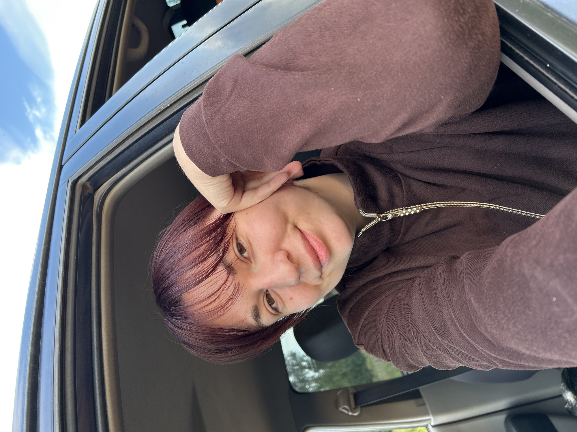 A young person with reddish-brown hair leans out of a car window, resting their head on their hand. They are wearing a brown zip-up hoodie. The car interior is visible, as is the exterior with a bright sky and clouds. The scene appears to be outdoors during the daytime, possibly in a rural or suburban area given the greenery visible through the window. The person's expression is relaxed and happy. The image was taken in Фоки, Russia.