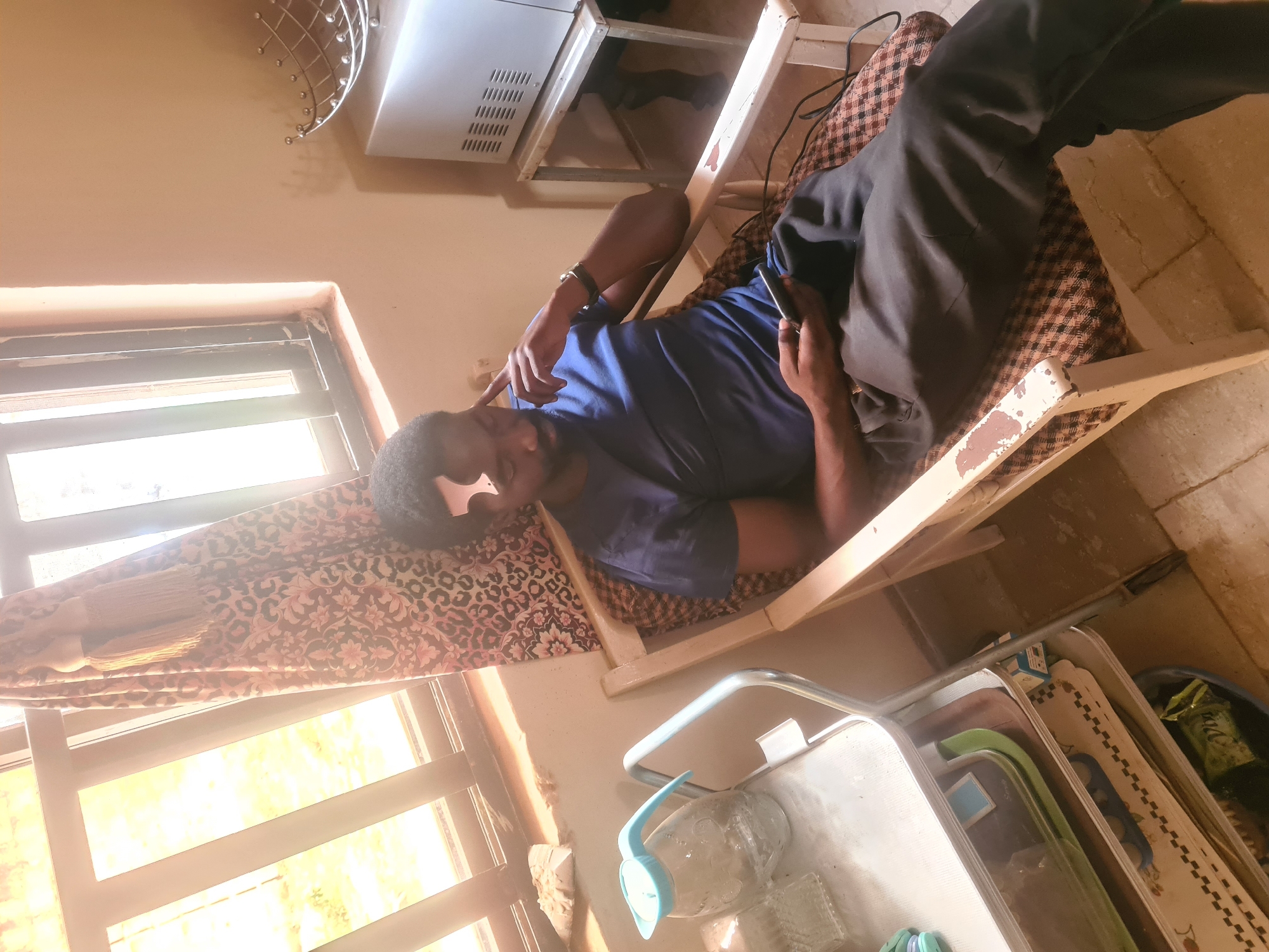 A man is depicted resting on a simple wooden cot with a patterned mattress in an indoor setting. He has light brown skin, short dark hair, and is wearing a blue t-shirt and dark trousers. He lies on his back with his head propped up, holding a dark object, likely a phone, in his left hand. His right hand is raised, with the index finger pointing upwards.
Bright light streams in from a window on the left, which has horizontal blinds and a patterned fabric (leopard print and floral) draped over it. The room has light-colored walls and a light-colored floor. Above the man's head, there's a metal wall decoration and a small white appliance, possibly a microwave, on a metal stand. Below the cot, a clear plastic water bottle with a blue spout is visible, along with a white utility cart containing various items like mats. The wooden frame of the cot shows signs of wear with peeling paint. The overall impression is one of relaxation in a modest living space during the daytime. There is no visible readable text.