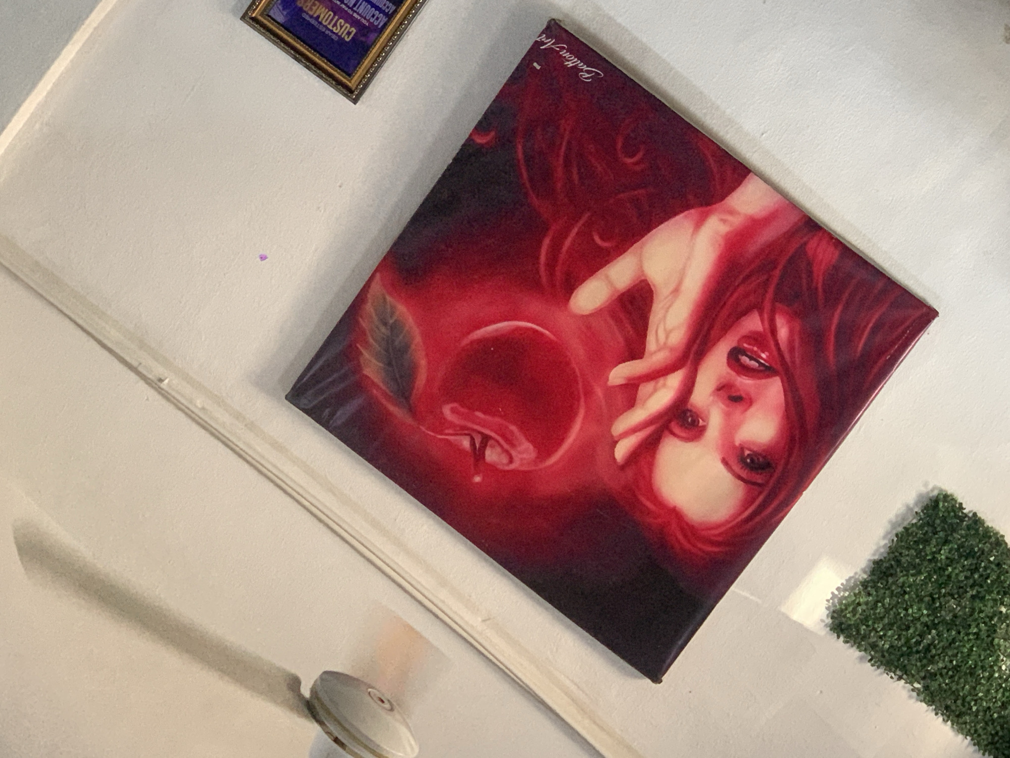 A painting of a woman with red hair and red lips, holding her hand up to her face, is displayed on a white wall. A red apple with a leaf is depicted above her. The painting is signed "Ballon Art" in the bottom right corner. In the upper left of the image, a portion of a white ceiling fan is visible, casting a shadow on the wall. To the right of the painting, a framed sign with the word "CUSTOMERS" and other text is partially visible. Below the painting, a small rectangular patch of artificial green turf is placed on the wall. The overall scene suggests an indoor setting, possibly a shop or a gallery, with the artwork being the focal point. The image was taken in Jalingo, Nigeria.