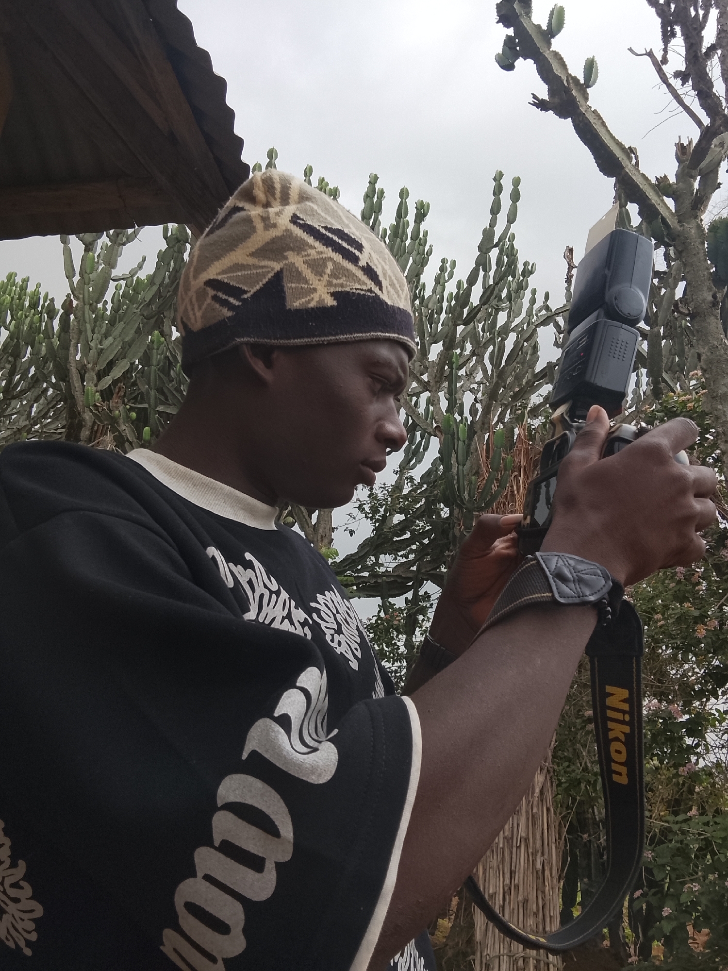 The image shows a man holding a Nikon camera with a flash attachment. He is wearing a patterned beanie and a black shirt with white text. A Nikon camera strap is visible on his arm. In the background, there are cactus-like plants and a corrugated metal roof structure. The location is in the city of Unknown, Unknown.
