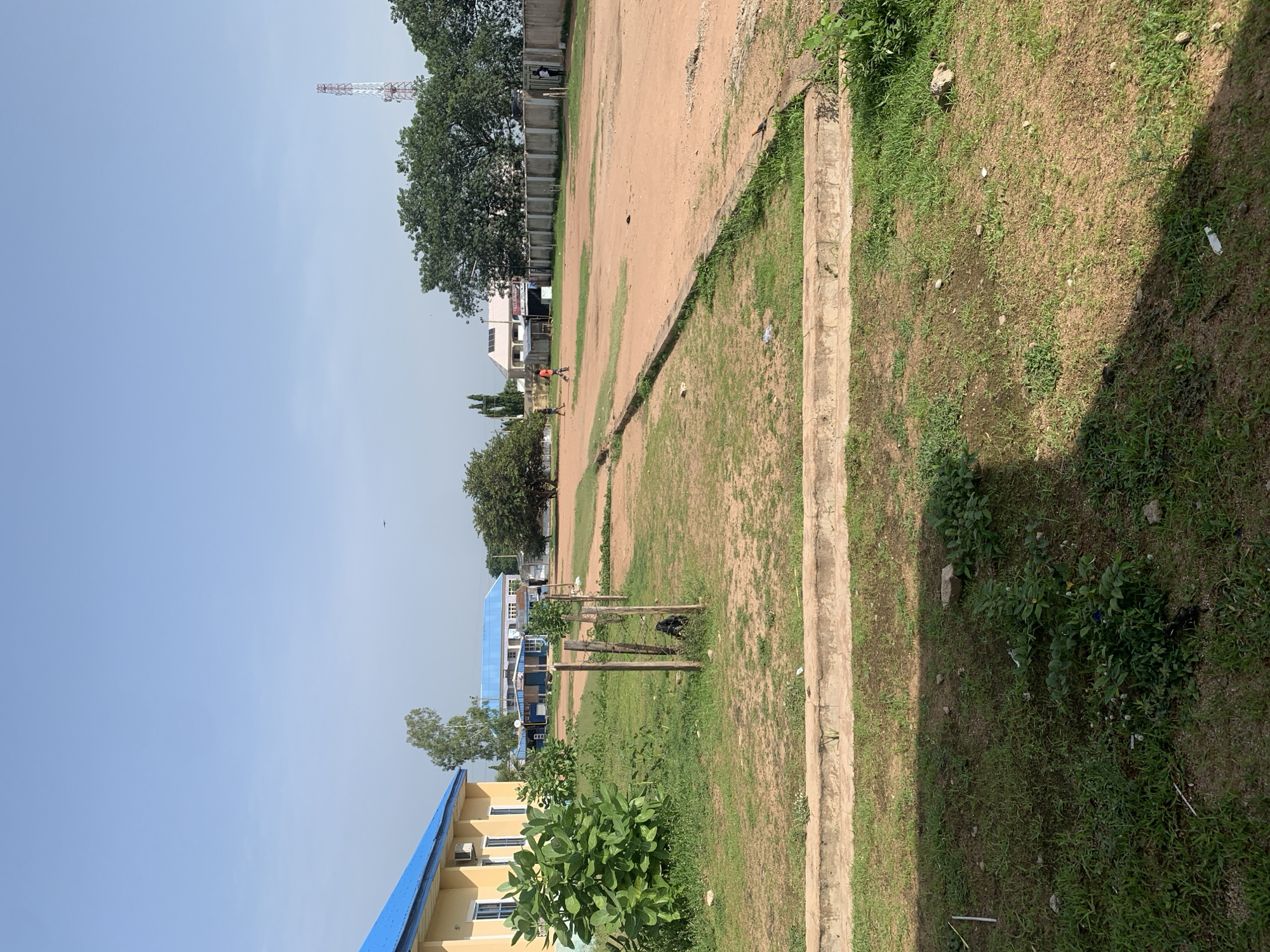 This is an outdoor, ground-level, eye-level, wide-angle shot of a public space in Jalingo, Nigeria. The scene features a building with a yellow facade and a blue roof on the left, overgrown green shrubs, a dirt field, and a concrete border in the foreground. In the mid-ground, a fenced-off area contains a dirt field where three individuals are walking. Beyond the field, there are trees, a white building with solar panels, and a telecommunications tower in the distance. The sky is clear and blue. The overall condition of the area appears to be dry and somewhat unkempt, with patches of dry grass and exposed soil.