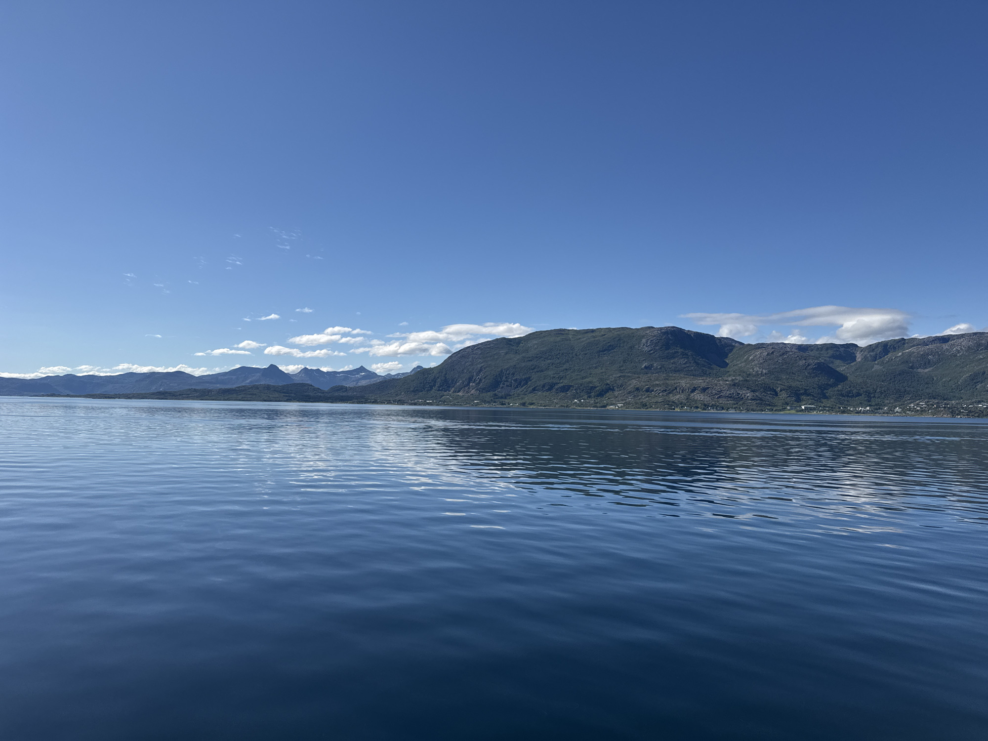The image depicts a serene seascape with mountains in the background, likely taken from a boat. The foreground is dominated by a vast expanse of calm, deep blue water, reflecting the clear blue sky above. Gentle ripples disturb the surface of the water, creating subtle patterns of light and shadow.
In the middle ground, a line of lush green mountains rises from the water's edge. The slopes of the mountains are covered in dense vegetation, and their rugged peaks are partially obscured by a few scattered white clouds. The outline of the mountain range stretches across the horizon, creating a dramatic backdrop to the tranquil water.
Along the base of the mountains, a small settlement is visible, with scattered houses and buildings nestled into the landscape. This suggests a coastal or fjord-like setting. The clear sky, bright sunlight, and calm waters indicate a pleasant, possibly summer day, with fair weather. The overall mood is peaceful and expansive, highlighting the natural beauty of the location. No people, objects, or text are clearly visible in the image. The location context of Lødingen, Norway, aligns with the visual cues of mountains meeting the sea in a scenic, northern European landscape.