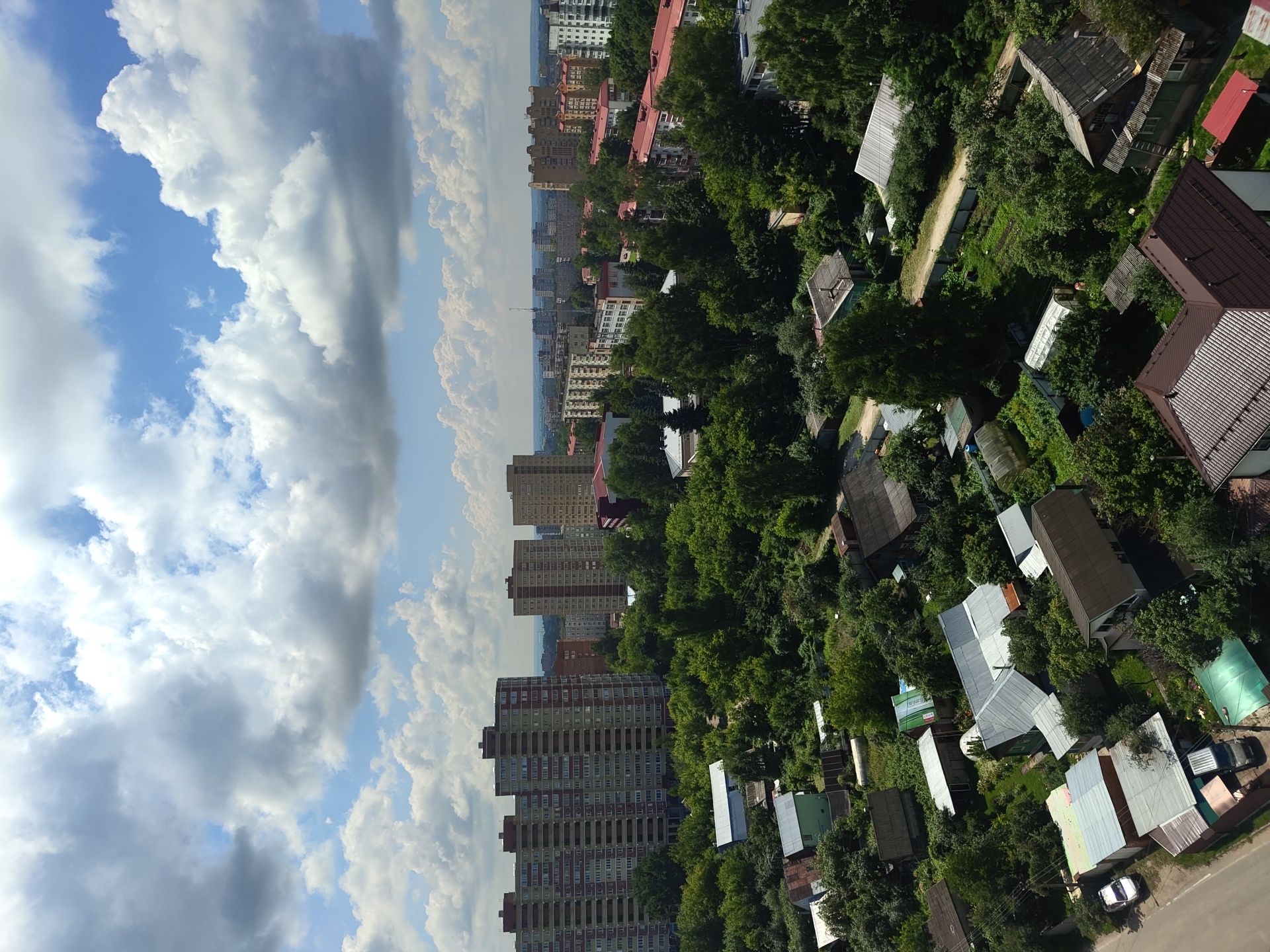 This is an aerial, eye-level, vertical, full shot of a city in Ramenskoye, Russia, under a bright, cloudy sky. In the distance, several tall, modern apartment buildings with many windows and red-brown accents are visible. In the middle ground, dense, green trees dominate the landscape, interspersed with smaller residential buildings, greenhouses, and garden plots. In the foreground, a few more houses with pitched roofs, some with metal sheeting, are visible, along with a paved road and parked vehicles. A white car and a dark van are parked on the road. The overall scene depicts a suburban area with a mix of apartment buildings and individual houses, surrounded by lush greenery.