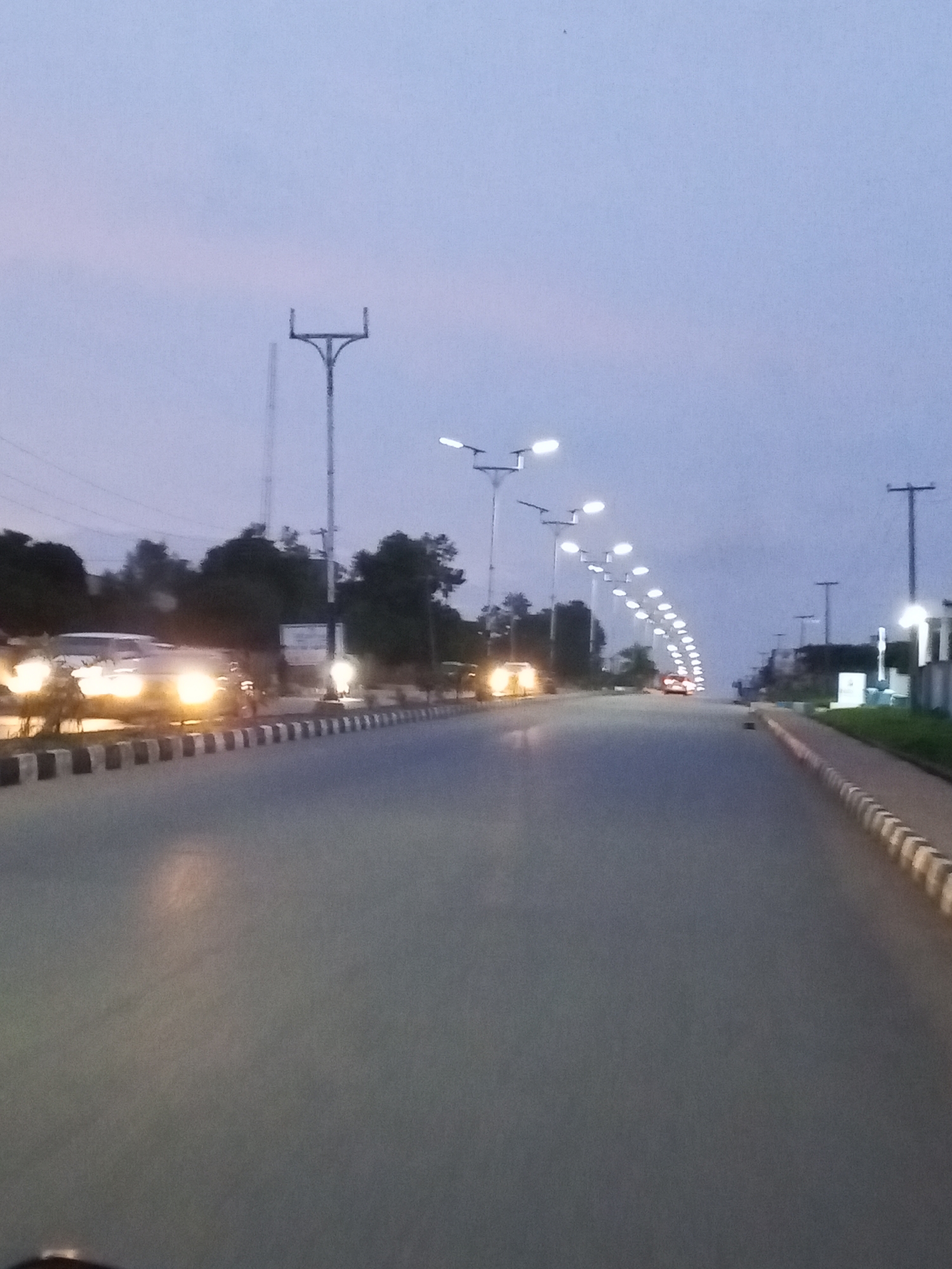The image depicts a dusk scene on a street in Jos, Nigeria. The sky is a pale blue, transitioning into twilight. A paved road stretches into the distance, lined on both sides with streetlights. Many of these streetlights are illuminated, casting a soft glow. On the left side of the road, several cars are visible, some with their headlights on, indicating they are in motion or have just stopped. The vehicles appear to be ordinary passenger cars. The road itself is a dark grey asphalt, with a white and black striped curb marking the edge of the pavement. Trees and some low vegetation can be seen behind the curbs on either side, forming a dark backdrop against the twilight sky. In the far distance, more streetlights form a glowing arc, suggesting a curved road or intersection. There are no visible people in the frame. The overall impression is that of a quiet, urban street at the end of the day.