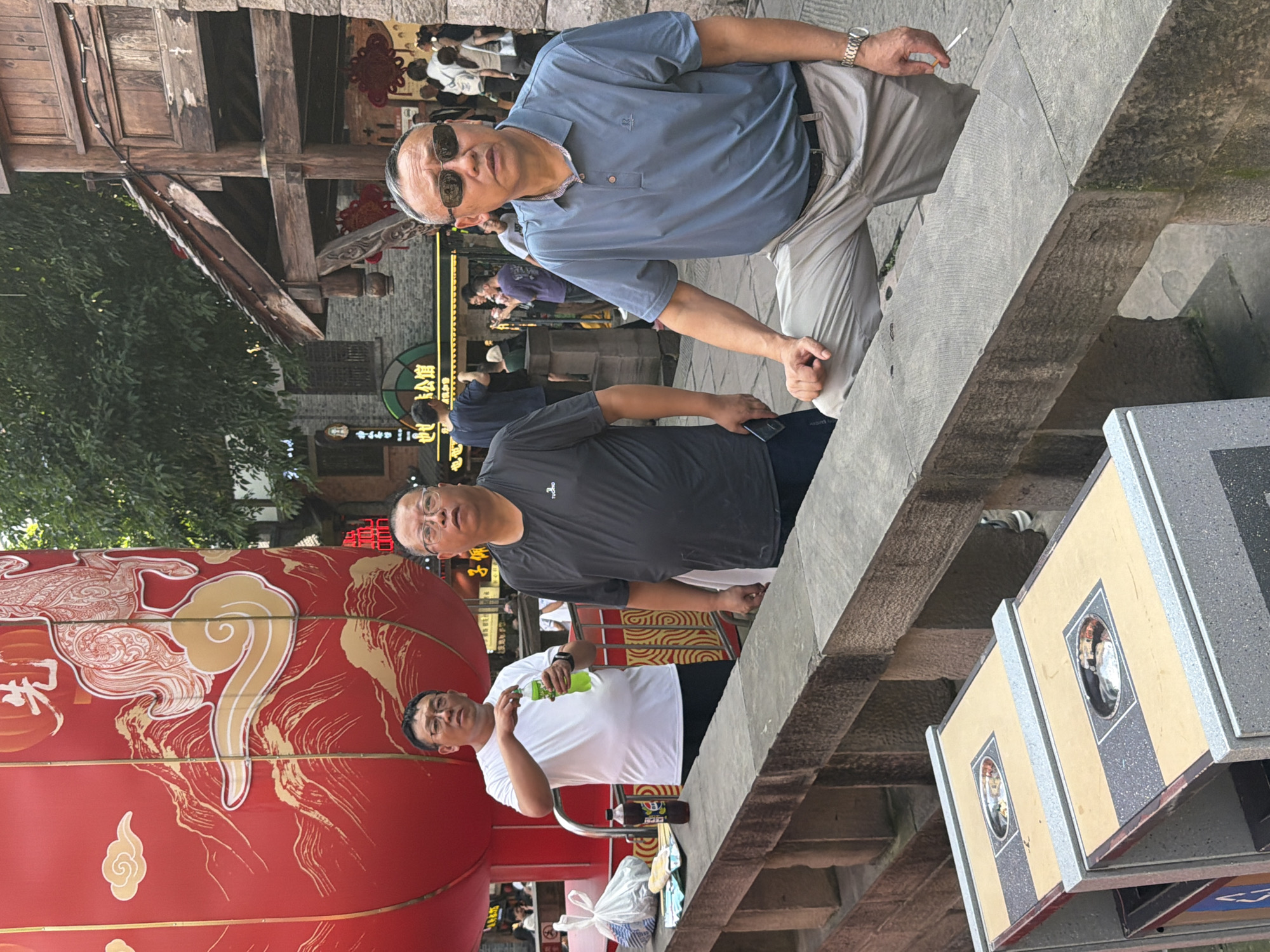 In a scene set in Yuzhong District, China, two men are captured in a candid moment, with another man partially visible. The primary subject, a man wearing a blue polo shirt and light-colored pants, stands with sunglasses and appears to be holding a cigarette. He looks towards the camera with a neutral expression. To his right, a man in a dark t-shirt and dark pants is also visible. Further down, a man in a white t-shirt is holding a green bottle and looking to his left. The setting suggests an outdoor area with traditional Chinese architecture, indicated by the wooden structures and a large red lantern with ornate gold designs. The time of day appears to be daytime, with natural light illuminating the scene. The overall atmosphere seems relaxed and casual, possibly a public space within a town or market. The background shows other people and buildings, suggesting a bustling environment. The visible text on the red lantern is in Chinese characters, but it is not clearly legible. There are also some display cases with framed images of people in traditional attire.