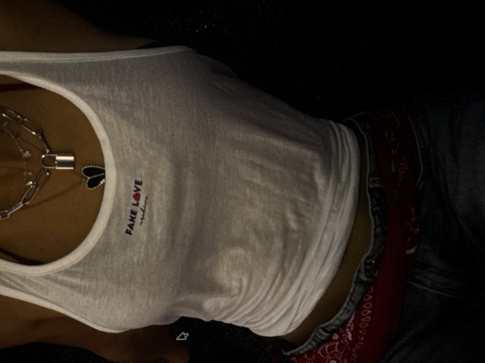 The image shows a close-up of a person's torso, lying down in a dimly lit environment. The person is wearing a white tank top with the text "FAKE LOVE" printed on it in red, with a small heart symbol replacing the 'o' in 'LOVE'. A silver chain necklace with a lock pendant and a black heart-shaped charm is visible around their neck. They are also wearing denim jeans and a red bandana peeking out from the waistline. The lighting suggests it might be nighttime or an indoor setting with minimal light. The overall mood is dark and mysterious. There are no discernible weather cues or specific location details, other than the provided context of Kuta Selatan, Indonesia.