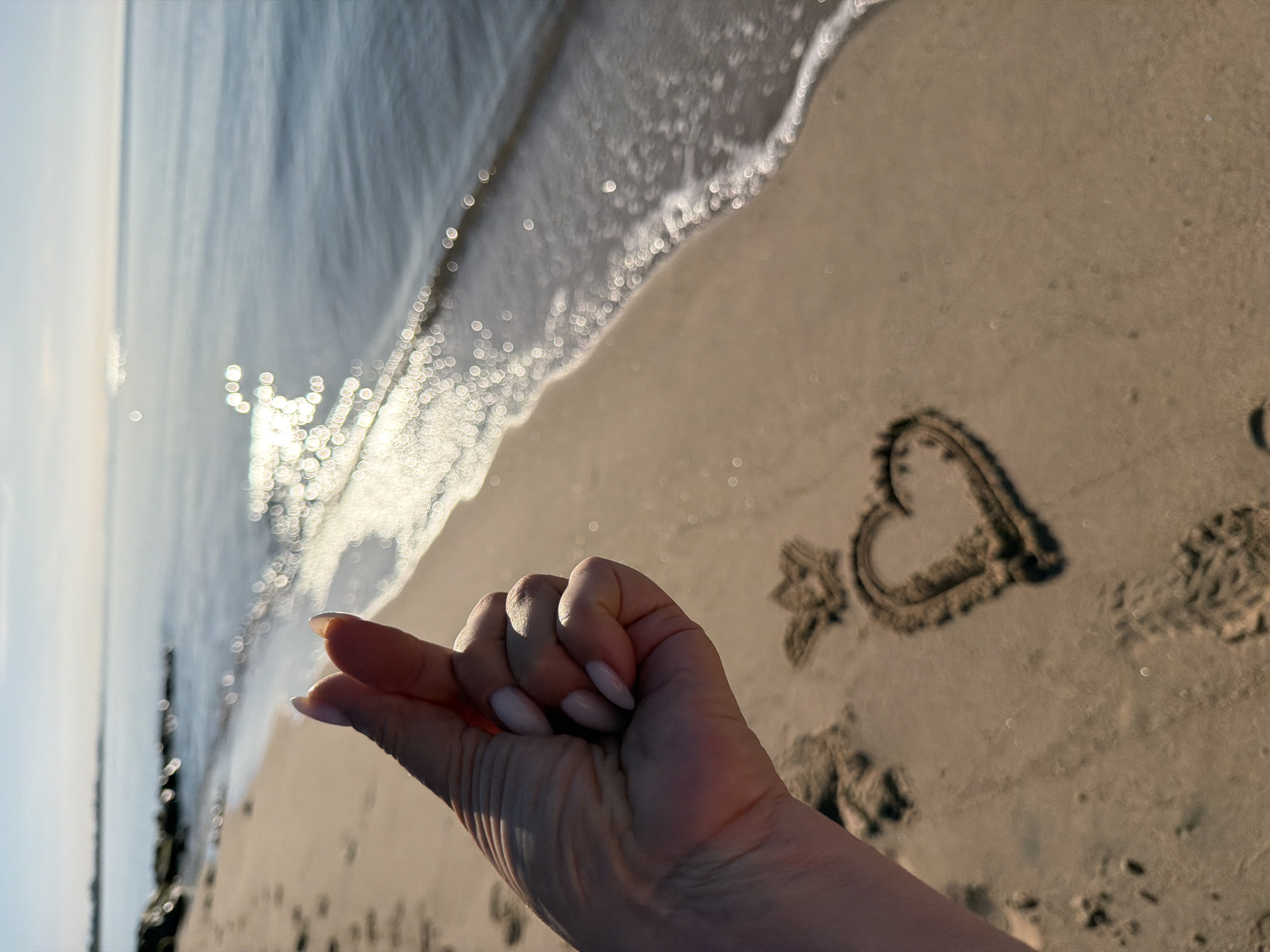The image shows a beach scene in Mont-roig del Camp, Spain, featuring a wave approaching the shore. In the foreground, a heart shape is drawn in the sand next to footprints. A person's hand with manicured fingernails is also visible, positioned near the sand drawing. The sun is reflected in the water. Wooden breakwaters are present near the water's edge.