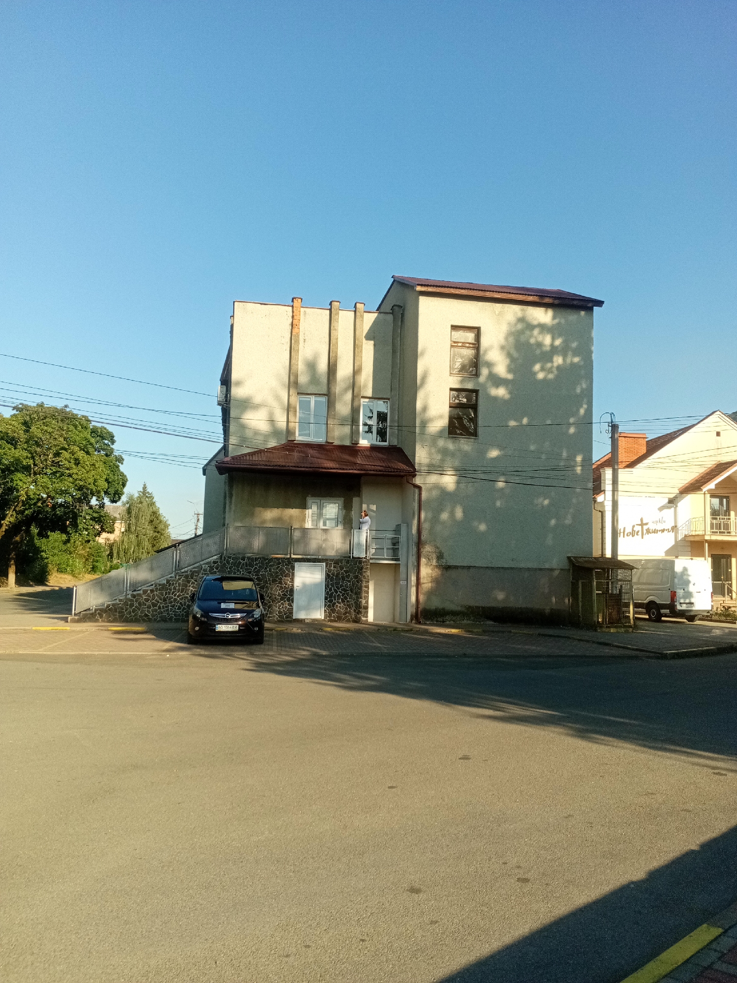 In Tiachiv, Ukraine, under a clear blue sky, a multi-story building with a light-colored facade stands prominently. The building features several windows of various sizes and a reddish-brown roof. A portion of the building's lower section is clad in stone. In front of the building, a dark-colored car is parked. To the right of the main building, another structure with a lighter facade and balconies is visible. Shadows from trees are cast across the building facades and the paved area in front. A small caged structure is situated near the side of the main building, and a white van is parked to the right, near the second building. A large green tree is located to the left of the main building. The scene appears to be a street or parking area in front of commercial or residential buildings.