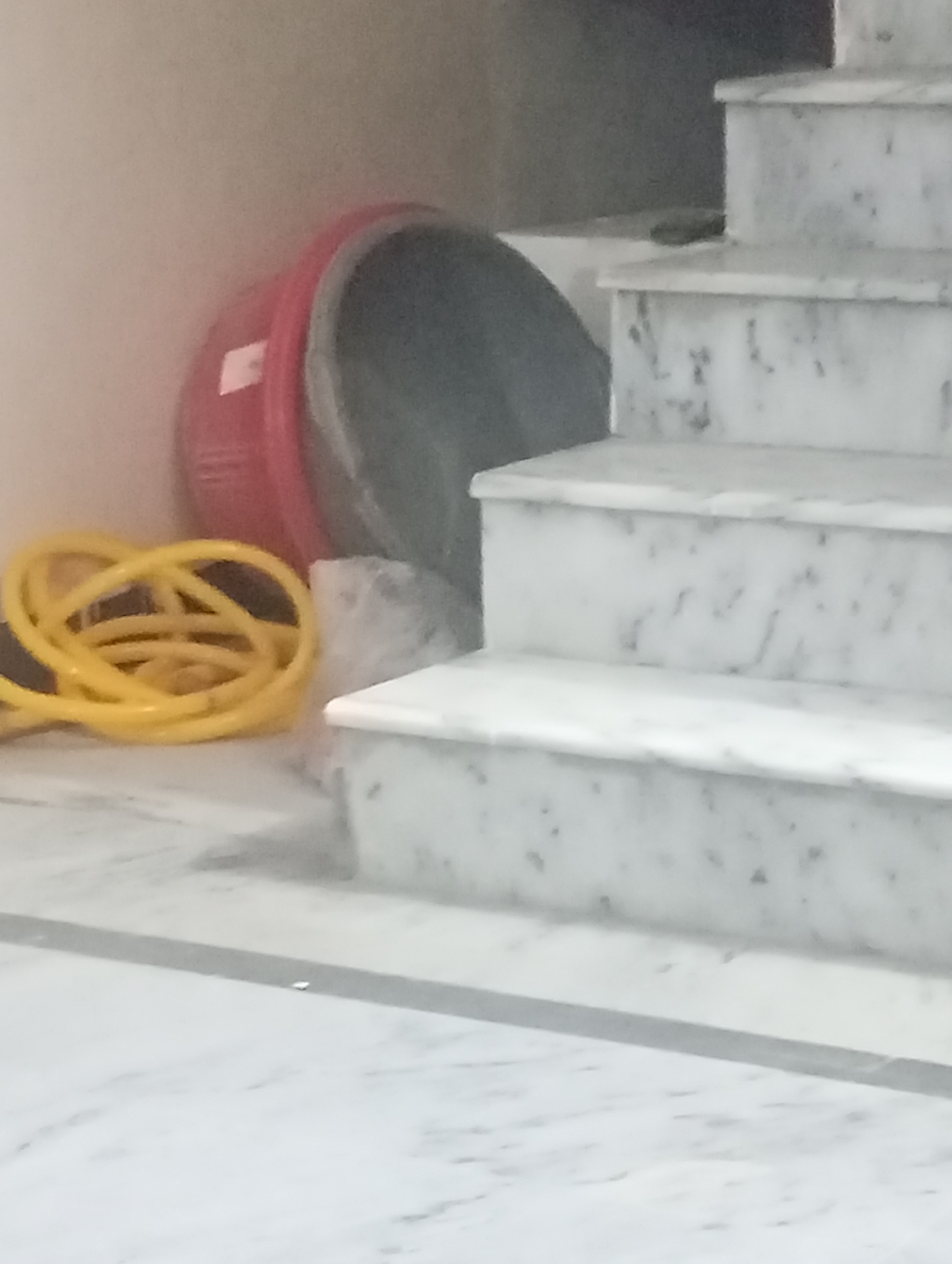 A red basin and a yellow coiled hose are on the floor next to marble stairs. The basin appears to be a concrete mixer.