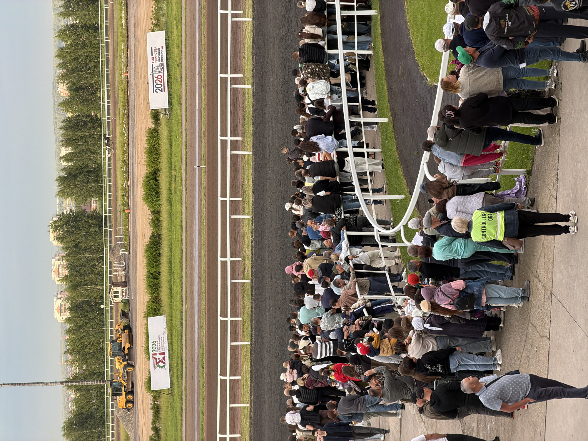 A wide shot captures an outdoor daytime scene at a horse racing track. A large crowd of people stands behind a white railing, observing the track. In the background, two yellow construction vehicles are parked near a small white building. A banner with Russian text and the year "2026" is displayed on the left. Another banner with similar text and "2026" is visible on the right. Beyond the track, trees and buildings form a distant landscape under an overcast sky. The foreground shows a paved area where some individuals are walking.