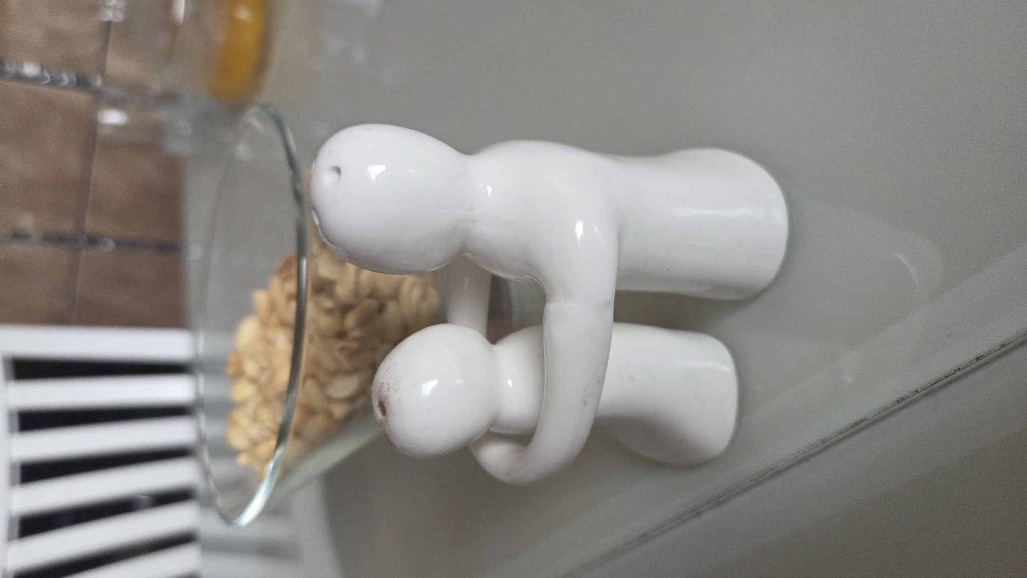 The image presents an indoor scene, likely a domestic setting such as a kitchen or dining area, in Chaikovsky, Russia. The central focus is two white ceramic figures, stylized to resemble human forms, arranged on a smooth, light-colored surface, possibly a countertop. These figures appear to be salt and pepper shakers, evidenced by small holes on the "head" of the upper figure.
One figure is positioned above the other, lying face down in a stance resembling a push-up or plank, supported by its ceramic "limbs." It has a glossy finish with minor smudges or dust visible on its surface. The second, smaller figure is placed beneath the first, with its rounded "head" and "torso" fitting snugly into the space created by the bent "arms" of the upper figure. This arrangement creates a whimsical and slightly acrobatic tableau, as if the top figure is carrying or supporting the one below.
To the left of these figures, a clear glass jar or bowl is partially visible, containing a light-brown, textured substance that resembles oats, granola, or a similar dried food mix. The background includes a blurry element with horizontal white slats, which could be part of a radiator cover or Venetian blinds. Further back, an indistinct yellow or orange object is faintly visible.
The lighting suggests an indoor setting, and no specific time of day can be determined. There is no visible text in the image. The composition appears to be a static display of decorative or functional items.