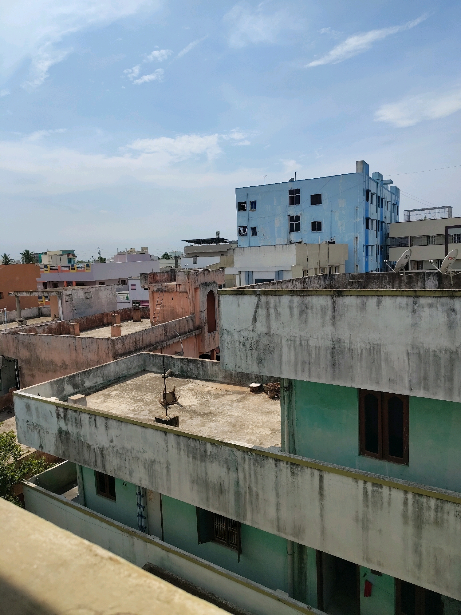 The image depicts a cityscape in Satyanarayana Puram, India, characterized by numerous low-rise buildings and a larger blue building in the background. The foreground is dominated by the rooftops of several buildings. The closest rooftop is a concrete terrace with a weathered, light-colored railing. To the left and behind this terrace, another rooftop is visible, also made of concrete and featuring a small, cylindrical structure, possibly a ventilation shaft or a water tank base, and a metal object resembling a chair or cart on a stand. Further back, more rooftops are visible, painted in various shades of pink, orange, and white, with some showing signs of age and wear. The blue building in the mid-ground has multiple floors and dark-colored windows. In the background, other buildings of varying heights and colors are scattered across the horizon, with some trees visible on the left. The sky is partly cloudy with a bright blue hue. The overall impression is of a densely populated urban area with a mix of old and new structures.