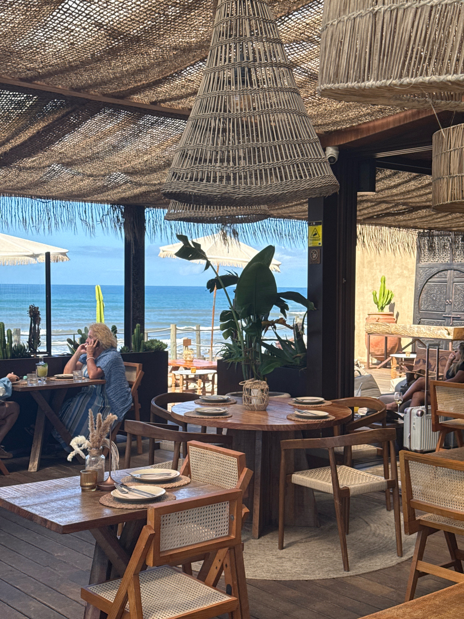 The image depicts an outdoor dining area with a view of the ocean. The restaurant has a rustic and bohemian aesthetic, with thatched roofing, woven light fixtures, and wooden furniture. Several tables are set for dining, some with plates, cutlery, and glasses.
In the foreground, a wooden table is set with two place settings, a small vase with dried flowers, and a glass votive candle holder. Two wooden chairs with woven seats are positioned at the table.
In the middle ground, a larger round wooden table is visible, also set for dining. A woman with blonde hair, wearing a blue and white striped dress, is seated at a table on the left, looking towards the ocean and speaking on her phone. She is seated at a wooden table with drinks and a glass. Behind her, there are umbrellas and a railing overlooking the beach.
In the background, the ocean is visible, with a clear blue sky above. There are some potted plants, including cacti, placed around the dining area, adding to the Mediterranean feel. The overall scene suggests a relaxed atmosphere at a seaside restaurant.
This establishment appears to be located in Vila Nova de Gaia, Portugal, a city known for its wineries and coastal charm, situated across the Douro River from Porto. The ocean view further supports this location.