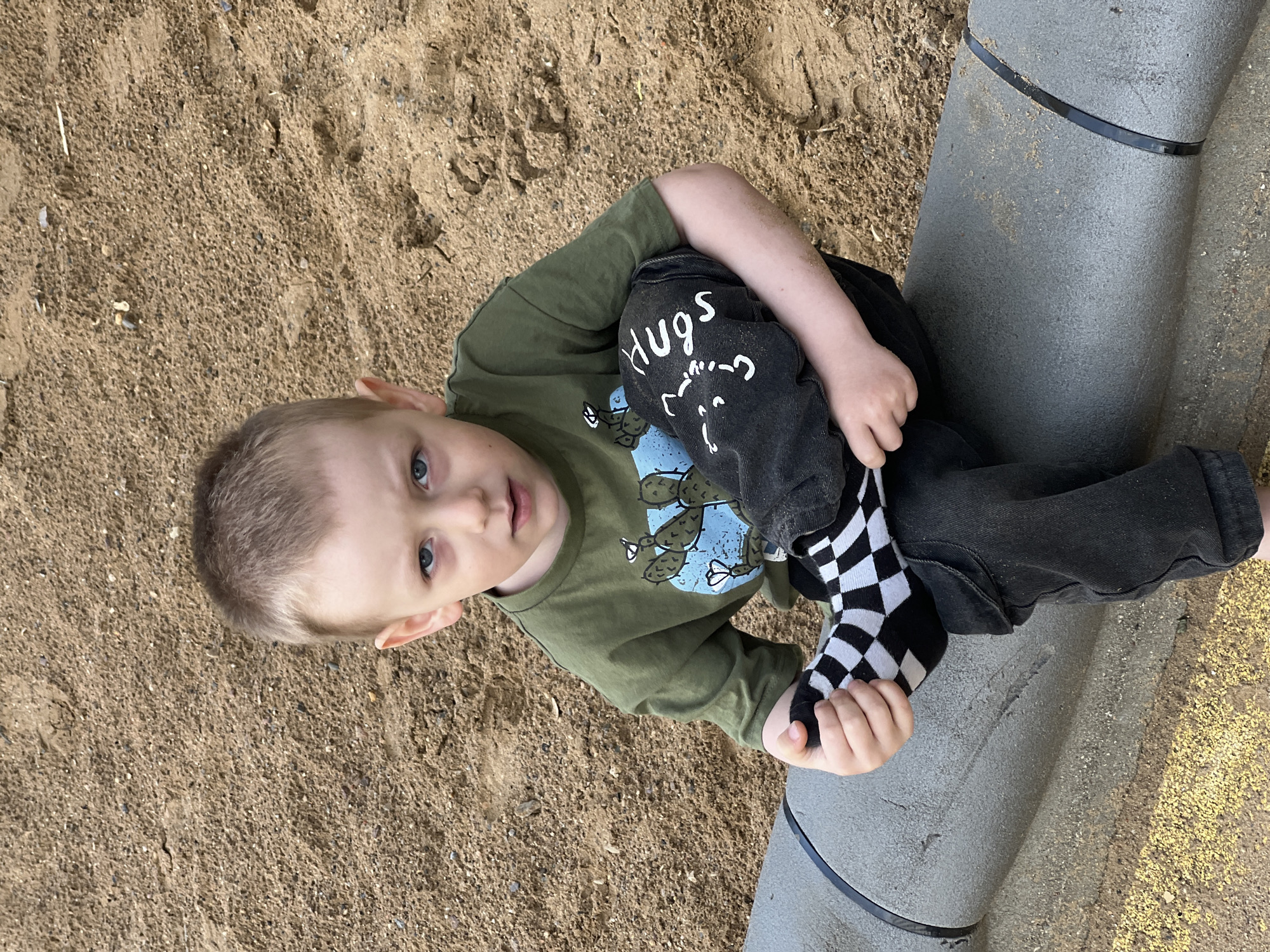 A young child sits on a large gray pipe, possibly part of a playground structure, located in an area with sand. The child is wearing a long-sleeved green shirt with a cactus design, black pants with the word "hugs" on one knee, and a black and white checkered sock on one foot. The environment appears to be a playground or park area. There is no other information provided about the location of Mytishchi, Russia.