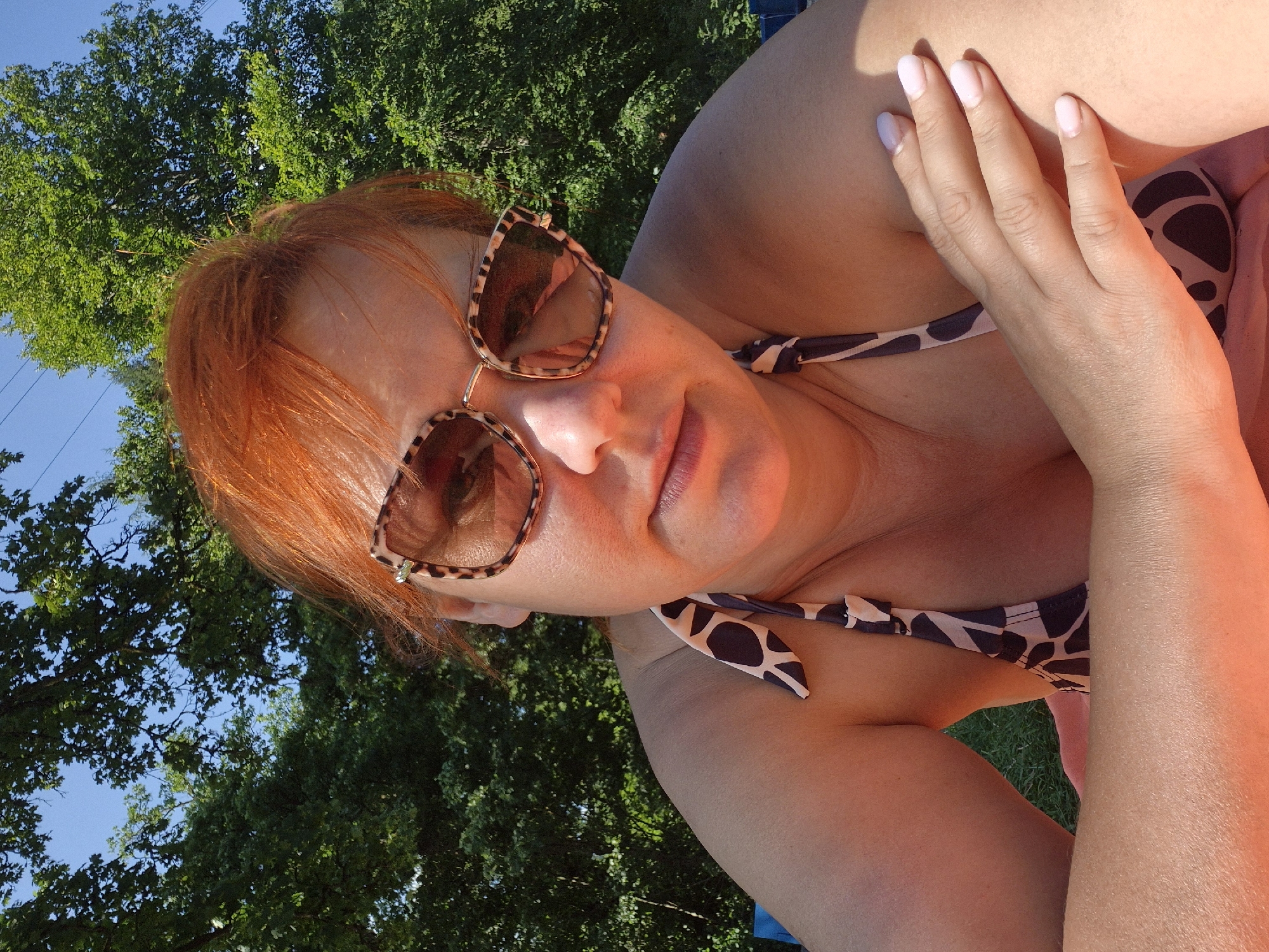 The image is a close-up, high-angle, outdoor selfie of a woman with reddish-orange hair. She is wearing large, tortoiseshell-patterned sunglasses and a bikini top with a brown and tan pattern. Her left arm is bent at the elbow, with her hand resting on her right shoulder. Her fingernails are painted a pale pinkish-lilac color. The background consists of lush green trees and a bright blue sky. The lighting suggests a sunny day. The woman appears to be in a relaxed, outdoor setting, possibly a park or garden, given the grass visible in the lower left corner. There are no visible landmarks that would definitively place this image in Schwäbisch Gmünd, Germany.