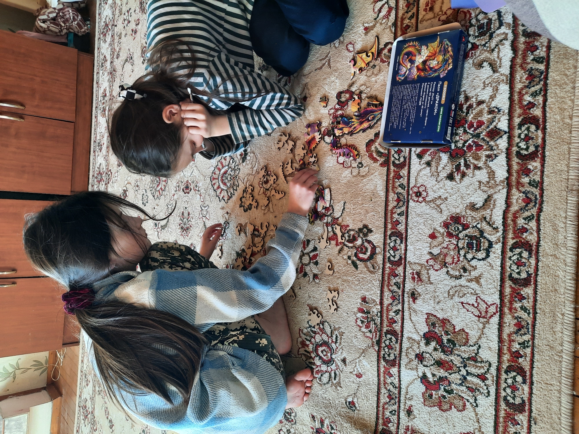 Two young girls are working on a jigsaw puzzle on a rug indoors. One girl wears a striped shirt and the other a blue and white plaid shirt. The puzzle box, featuring a dragon, rests on the rug near scattered puzzle pieces. The scene appears to be in a home, possibly in Архангельское, Russia given the Cyrillic writing on the puzzle box. A wooden cabinet is visible in the background.