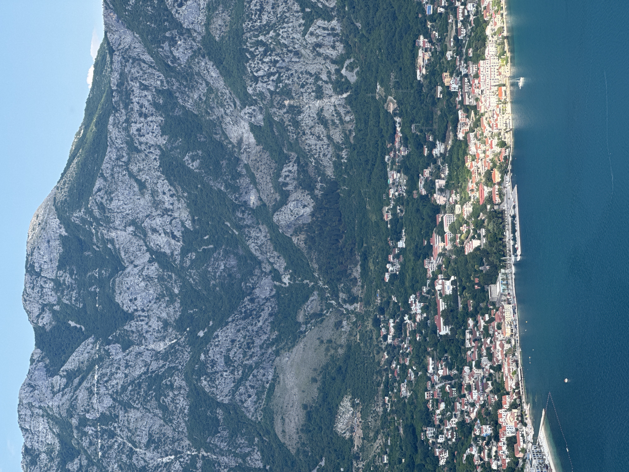 A high-angle aerial view captures the coastal town of Štrp, Montenegro, nestled at the foot of a towering, rocky mountain range. The deep blue waters of the bay occupy the foreground, with a few small boats and buoys dotting its surface. The town itself is a dense collection of buildings with red-tiled roofs, stretching along the coastline and climbing up the lower slopes of the mountain. Greenery is abundant, with trees surrounding and interspersing the urban landscape. The mountain behind the town is rugged and sparsely vegetated, with visible rock faces and patches of dark green trees. The sky is a clear, pale blue. The image appears to be taken on a sunny day.