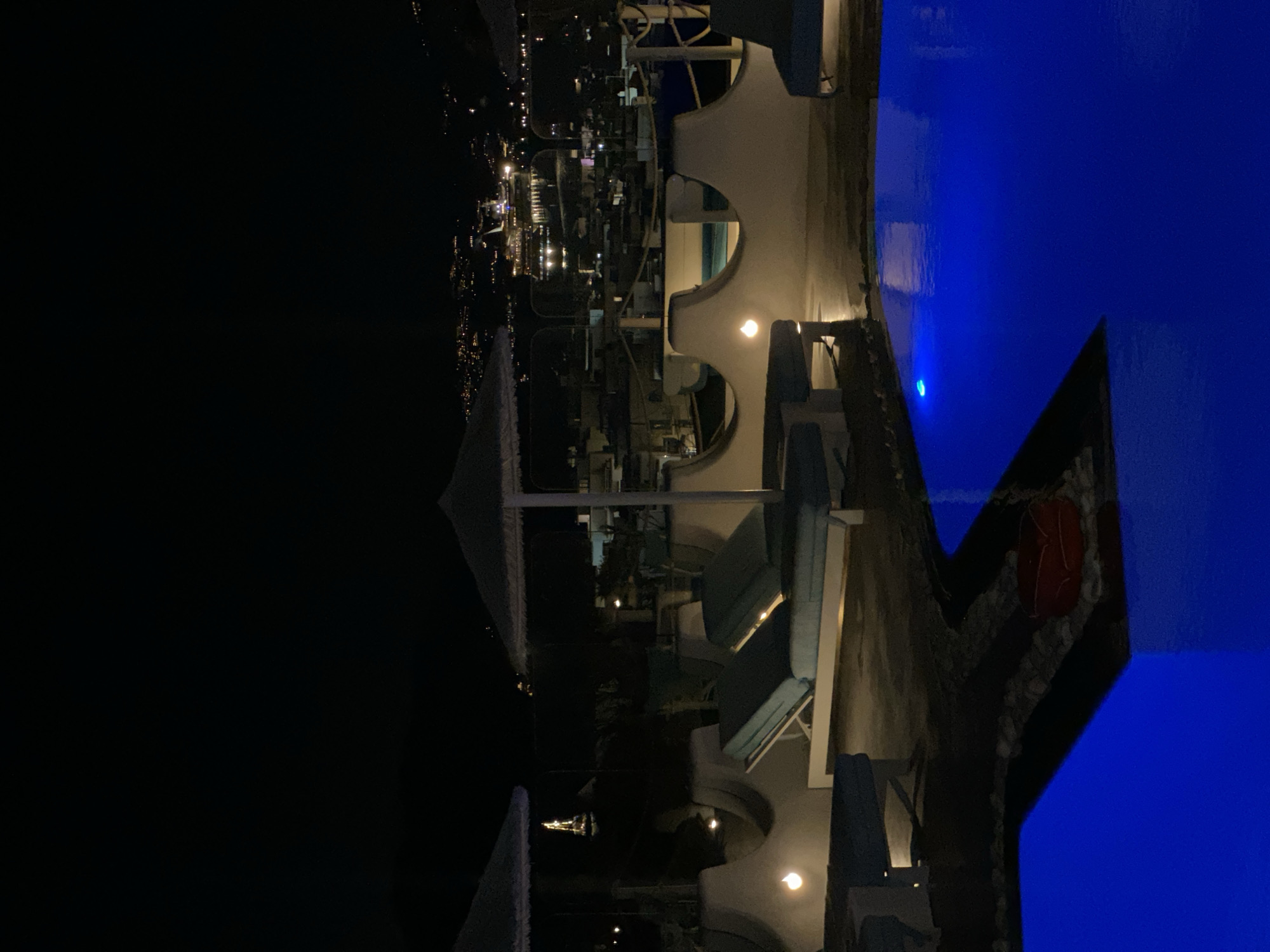This is a nighttime shot of a rooftop pool area with a view of Mykonos town and a cruise ship in the distance. The foreground shows the bright blue water of the pool, with some lounge chairs and umbrellas visible on the surrounding deck. In the middle ground, there are more lounge chairs and low, curved walls with small lights. The background reveals the lights of Mykonos town and a large illuminated cruise ship docked in the harbor. The sky is dark and clear, with a few distant lights suggesting a faint skyline. The overall atmosphere is serene and luxurious, typical of a high-end resort in Mykonos.