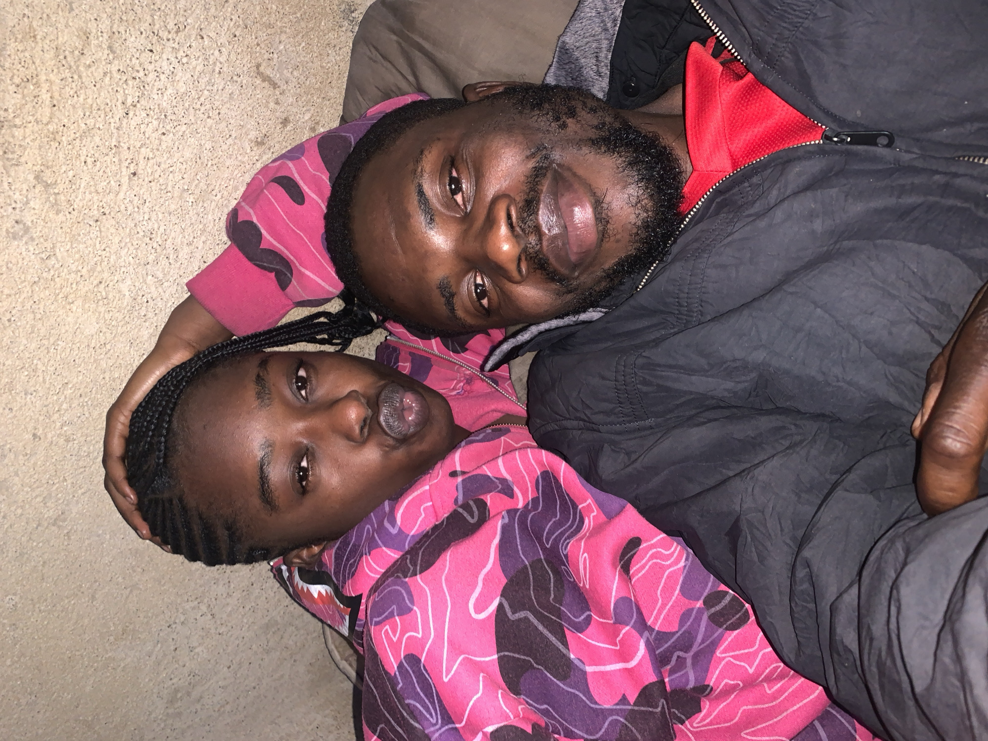 The image shows a close-up selfie of two people, a young woman and a man, who appear to be lying down. The woman, on the left, has dark skin, braided hair, and is wearing a pink and purple camouflage patterned hoodie. She is leaning her head on her hand and making a kissing face. The man, on the right, has dark skin, a beard, and is wearing a black jacket over a red shirt. He is looking towards the camera with a slight smile. They are positioned against a textured, neutral-colored wall, which could be a stucco or concrete surface. The lighting appears to be dim, possibly from an artificial source, casting soft shadows. The overall impression is an informal, personal photograph taken in a domestic or private setting. It is not possible to determine the specific location within the city of Rafin Par, Nigeria, from the image alone.