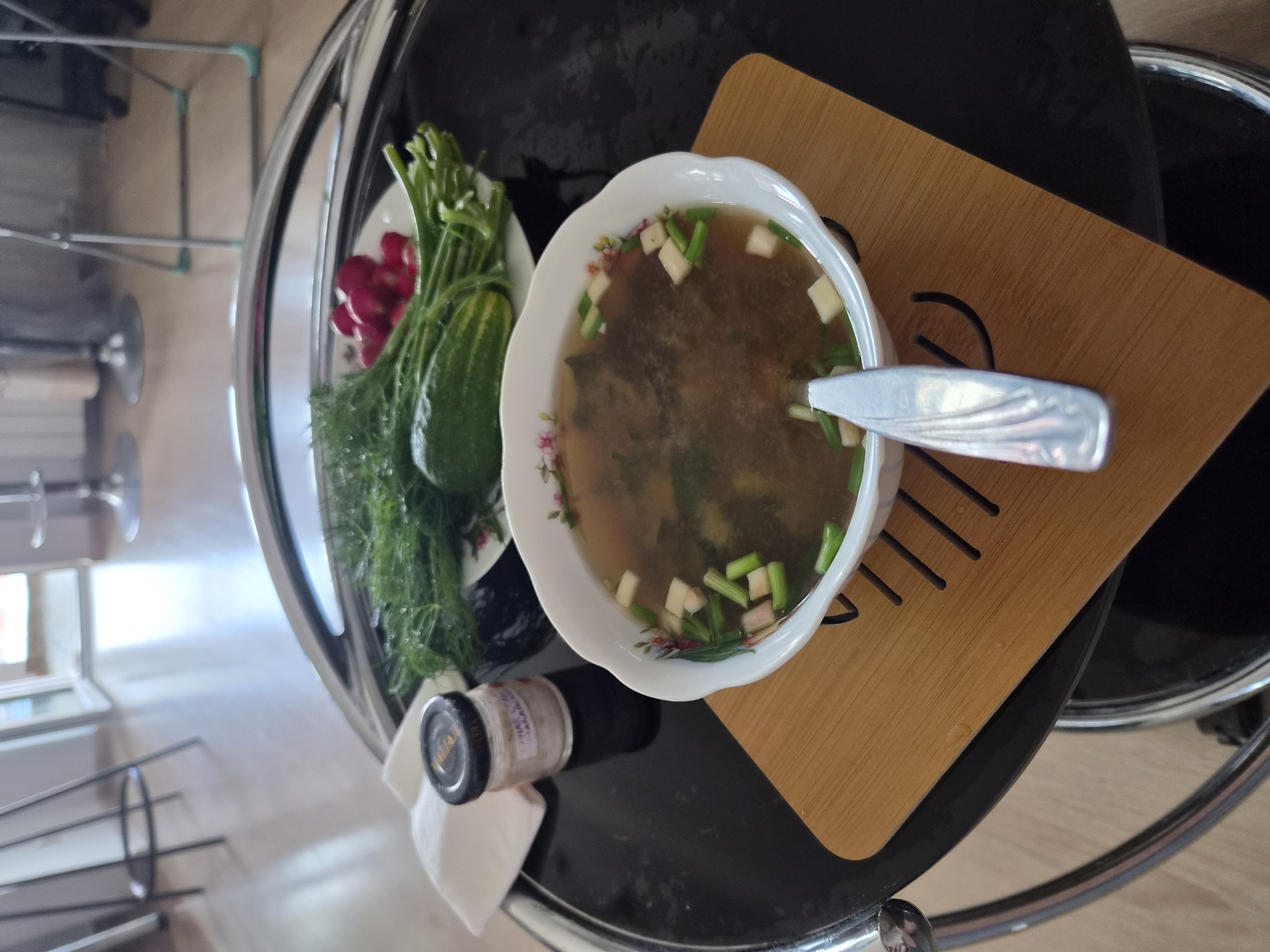 A bowl of soup garnished with chopped green onions sits on a wooden placemat on a black round table. A plate of dill, cucumbers, and radishes is to the left of the soup. A small jar of pink salt is next to the plate. The table is in a room with a wooden floor and chairs in the background. The scene appears to be indoors, possibly in a residential setting or a cafe, in Vladivostok, Russia.