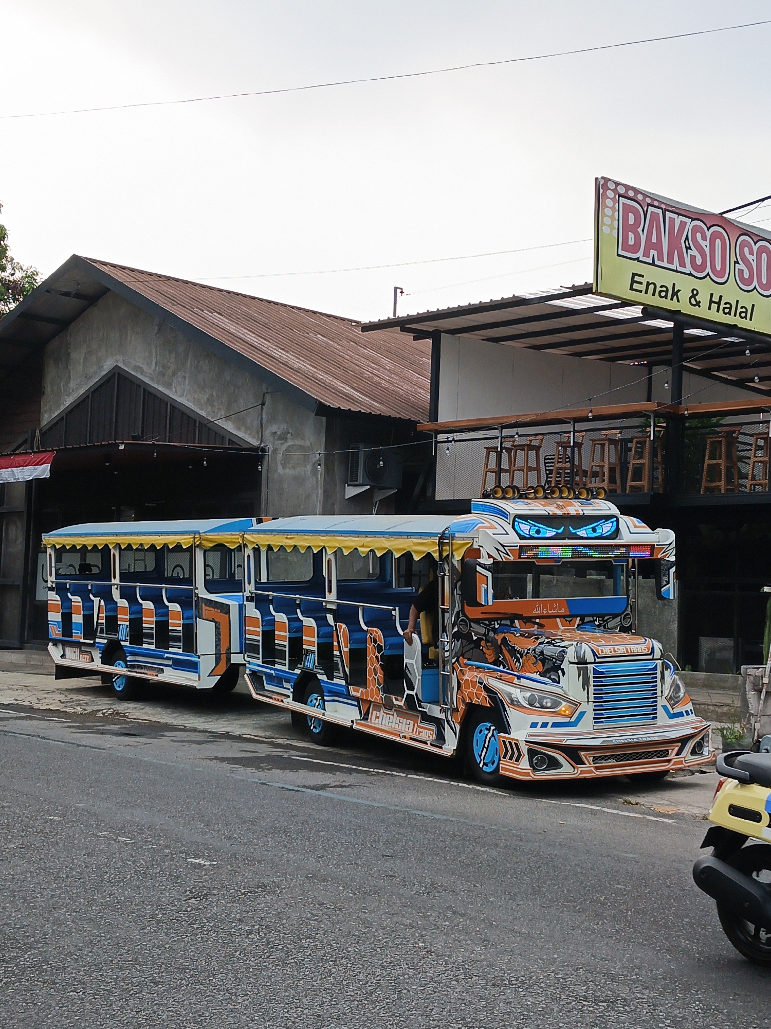 An eye-level, slightly low-angle, full shot captures a vibrant, custom-built tourist bus, resembling a train, parked on a paved road in Klaten, Indonesia. The bus is painted in a striking combination of blue, orange, and white, with yellow accents on its canopy. It features an elaborate design with what appears to be an animal motif, possibly giraffes, adorning its front. The bus has multiple rows of open-air seating with blue upholstery and blue wheel rims. The front of the bus is designed with aggressive, stylized headlights that resemble eyes.
Behind the bus, a building with a corrugated metal roof and concrete walls is visible. A sign with the text "BAKSO SO" and "Enak & Halal" is prominently displayed above a covered outdoor seating area with wooden chairs and tables. The environment suggests a commercial area, likely a restaurant or food stall. To the right of the bus, the rear of a yellow scooter is partially visible, indicating its presence on the road. The sky is overcast, suggesting a daytime scene. The overall impression is one of a lively, tourist-oriented location within the city of Klaten.