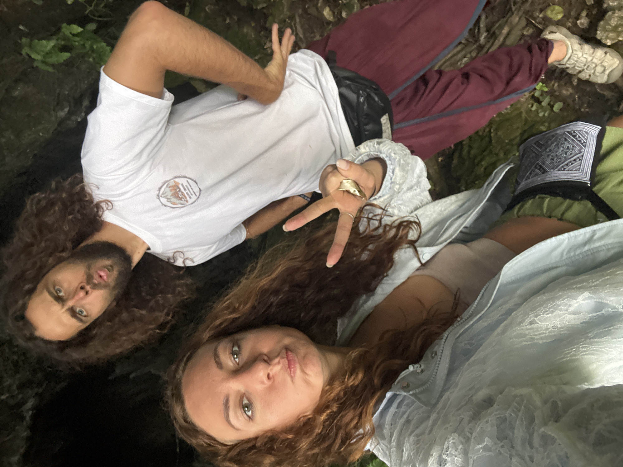 The image shows two individuals, a woman and a man, standing in what appears to be a natural, possibly cave-like, outdoor environment. The woman is in the foreground on the left, taking a selfie. She has long, curly brown hair and is wearing a light blue lace jacket over a beige top. Her facial expression is a pout, and she is holding up two fingers in a peace sign with her right hand, which has a large gold ring on her index finger.
The man is in the background on the right, looking towards the camera with a surprised or playful expression. He has long, curly brown hair and a beard. He is wearing a white t-shirt with a circular logo on the left chest and maroon pants. A black fanny pack is worn around his waist. His left hand is on his hip, and his right arm is bent, with his hand gesturing outwards.
The environment appears to be dimly lit with dark, rocky formations visible behind the man. There is some greenery and moss on the ground and rocks, suggesting a humid, natural setting. The specific location within Mai Chau, Vietnam, is not discernible from the visual information in the image.